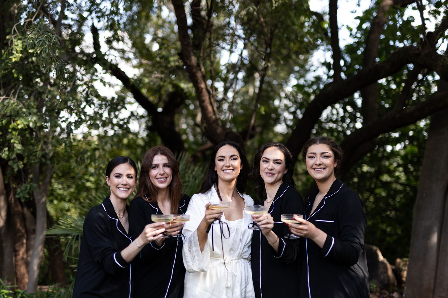 Bridal party in coordinated outfits pose together with champagne glasses during outdoor celebration.