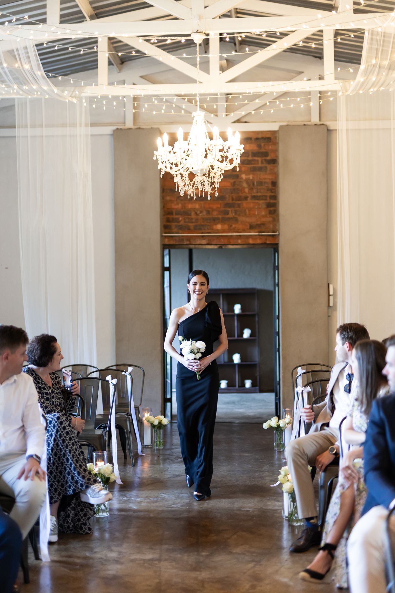 Bridesmaid in navy dress walks down aisle under crystal chandelier in industrial-chic wedding venue.