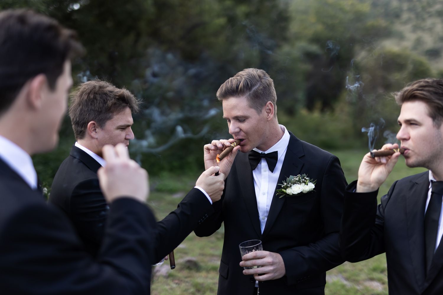 Wedding party members in formal attire smoking cigars at an outdoor gathering.
