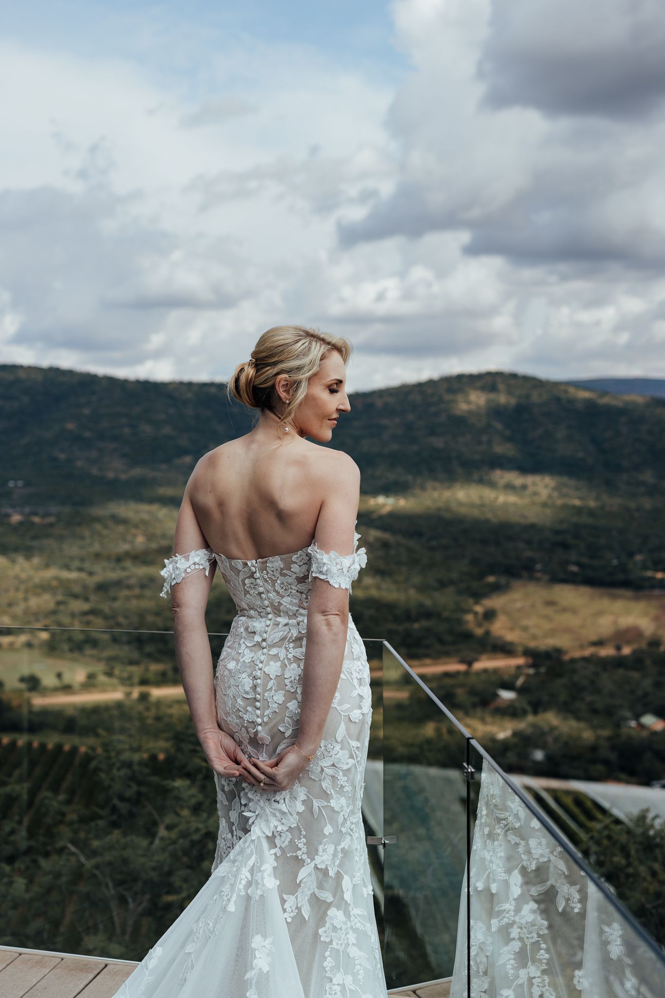 Bride in off-shoulder lace gown stands on balcony overlooking mountain landscape.