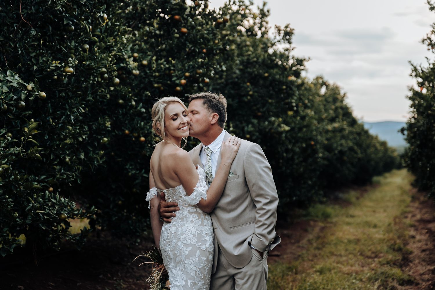 Romantic wedding couple embraces in citrus grove during sunset photoshoot.