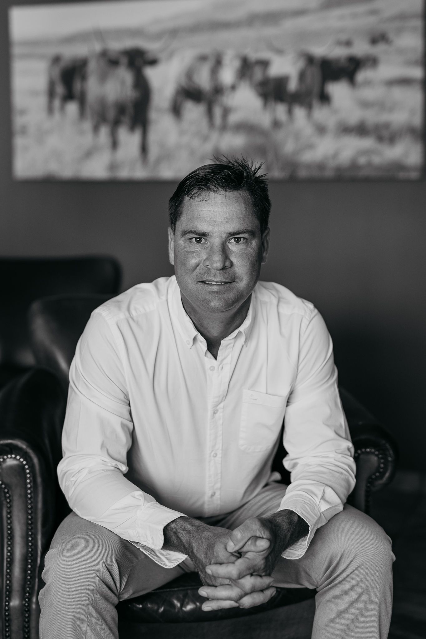 Black and white portrait of person seated in leather chair with horse artwork backdrop.