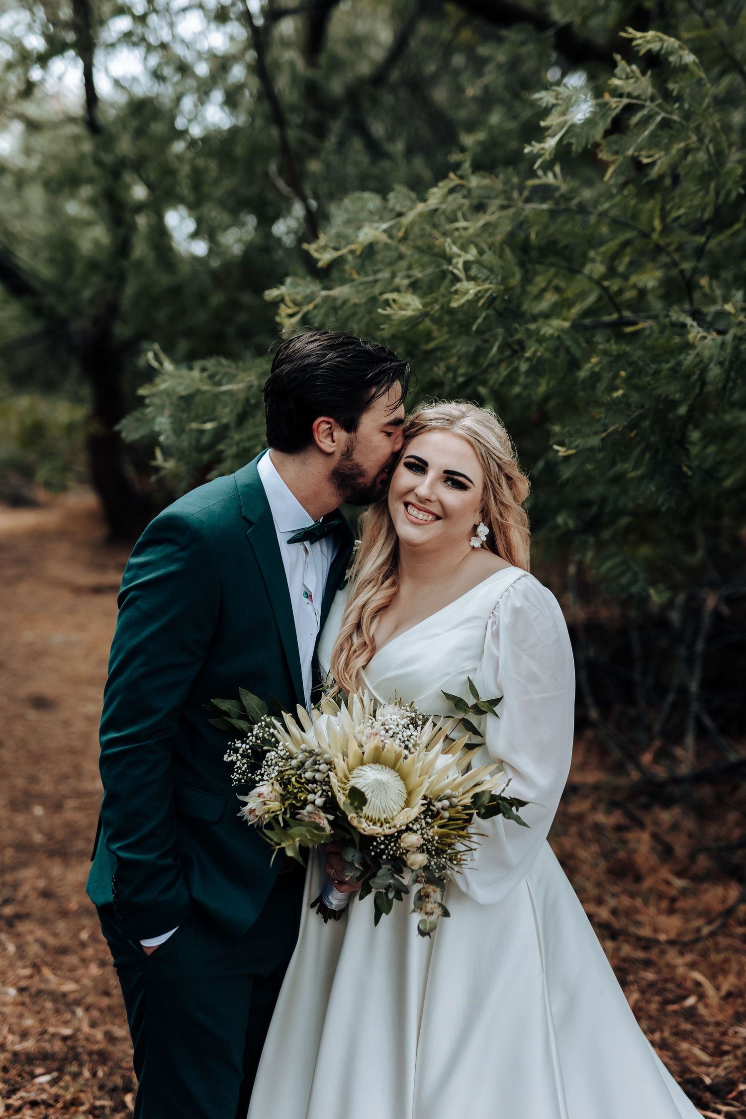 Forest wedding portrait with couple sharing sweet moment and bride holding elegant white bouquet.