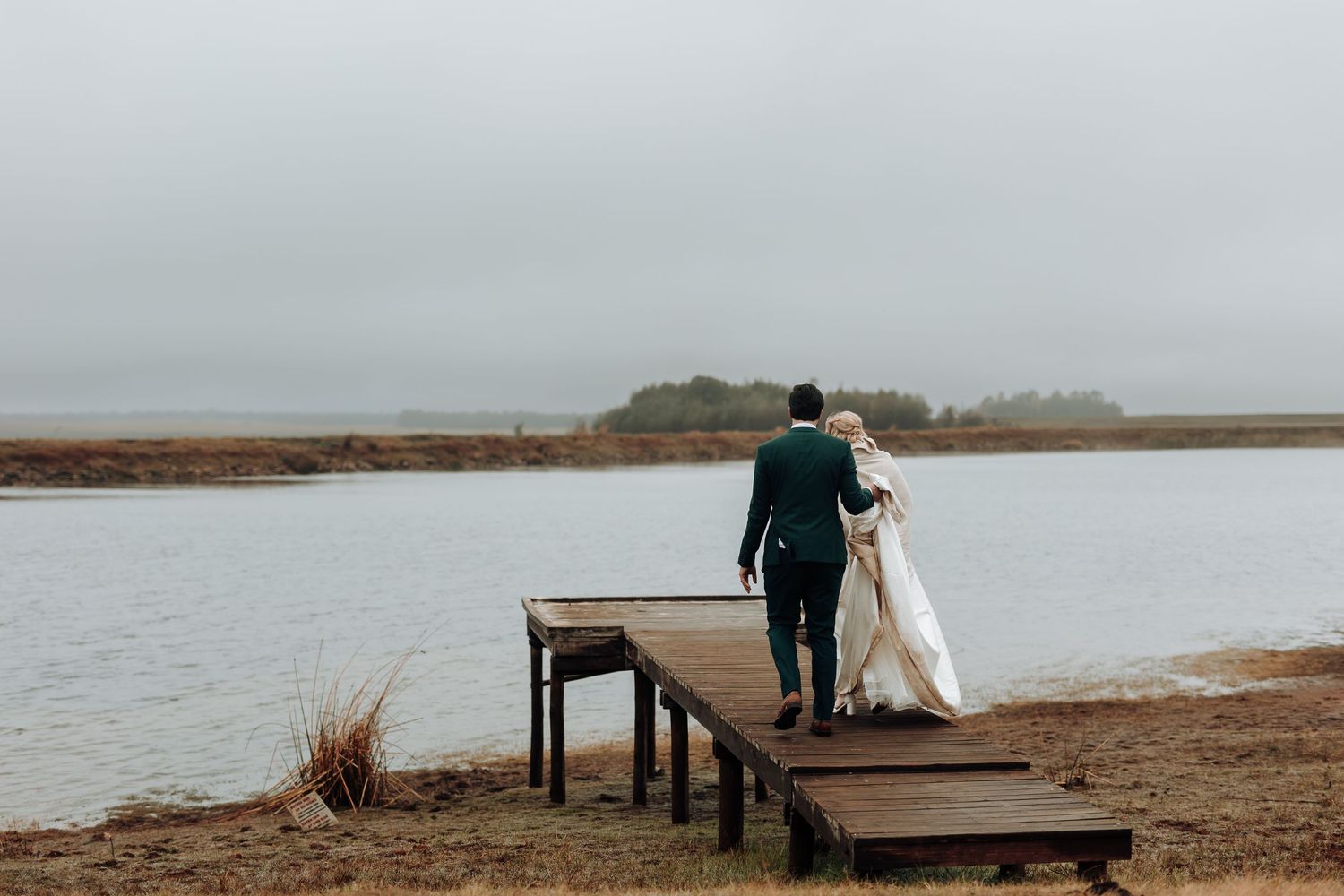 Couple walks hand in hand on wooden pier overlooking tranquil water.