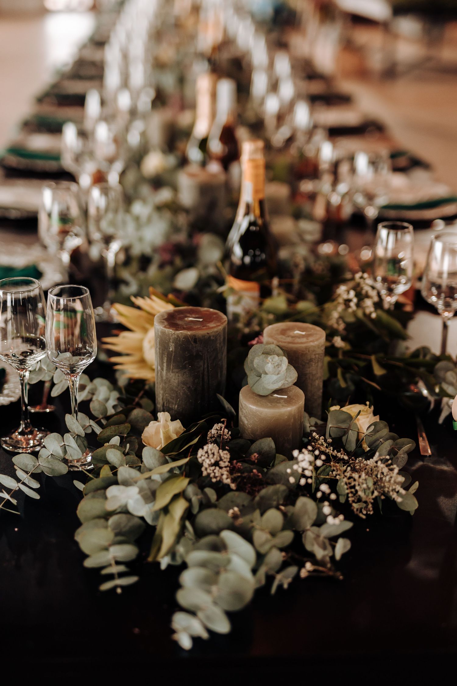 Moody wedding table centerpiece with candles, eucalyptus, and wine glasses on dark tablecloth.