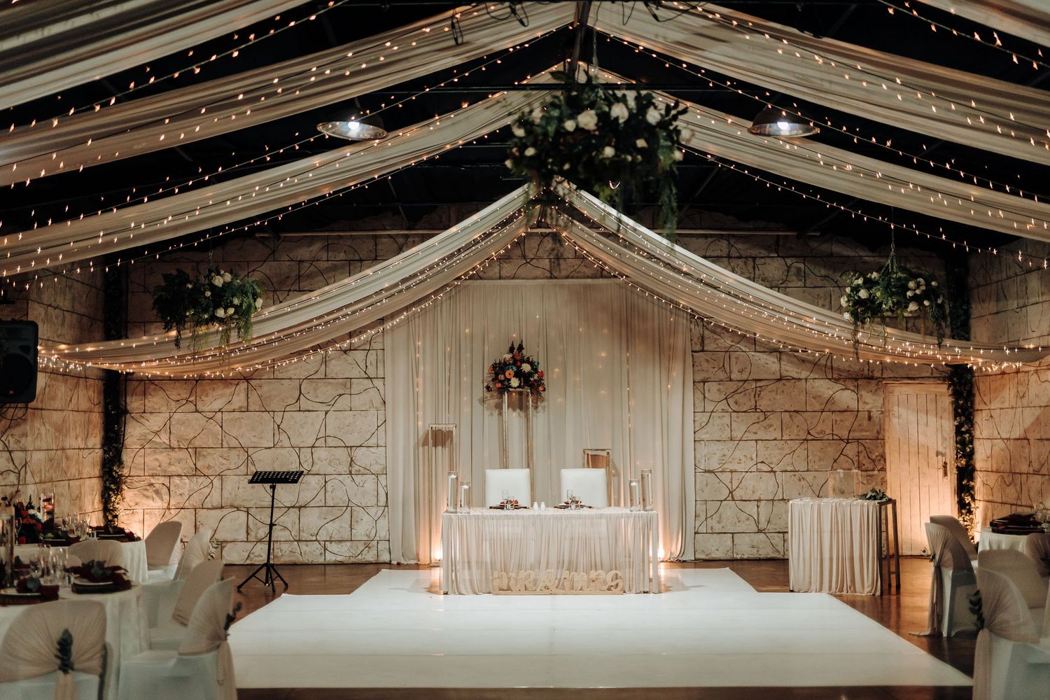 Wedding reception setup with white draping, string lights, and elegant head table in a rustic stone venue.