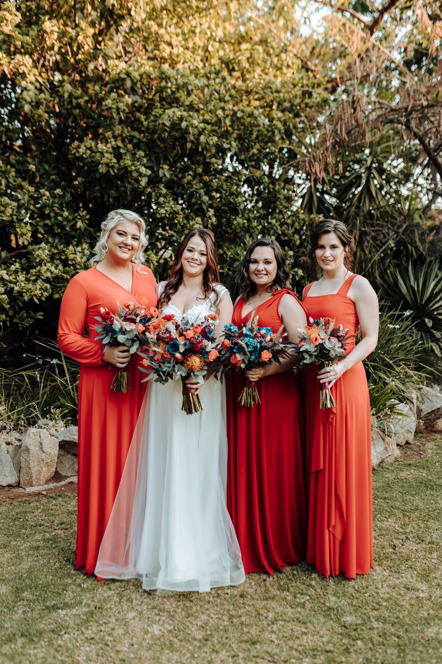 Bridal party in coral and red dresses pose with bouquets against lush garden backdrop.