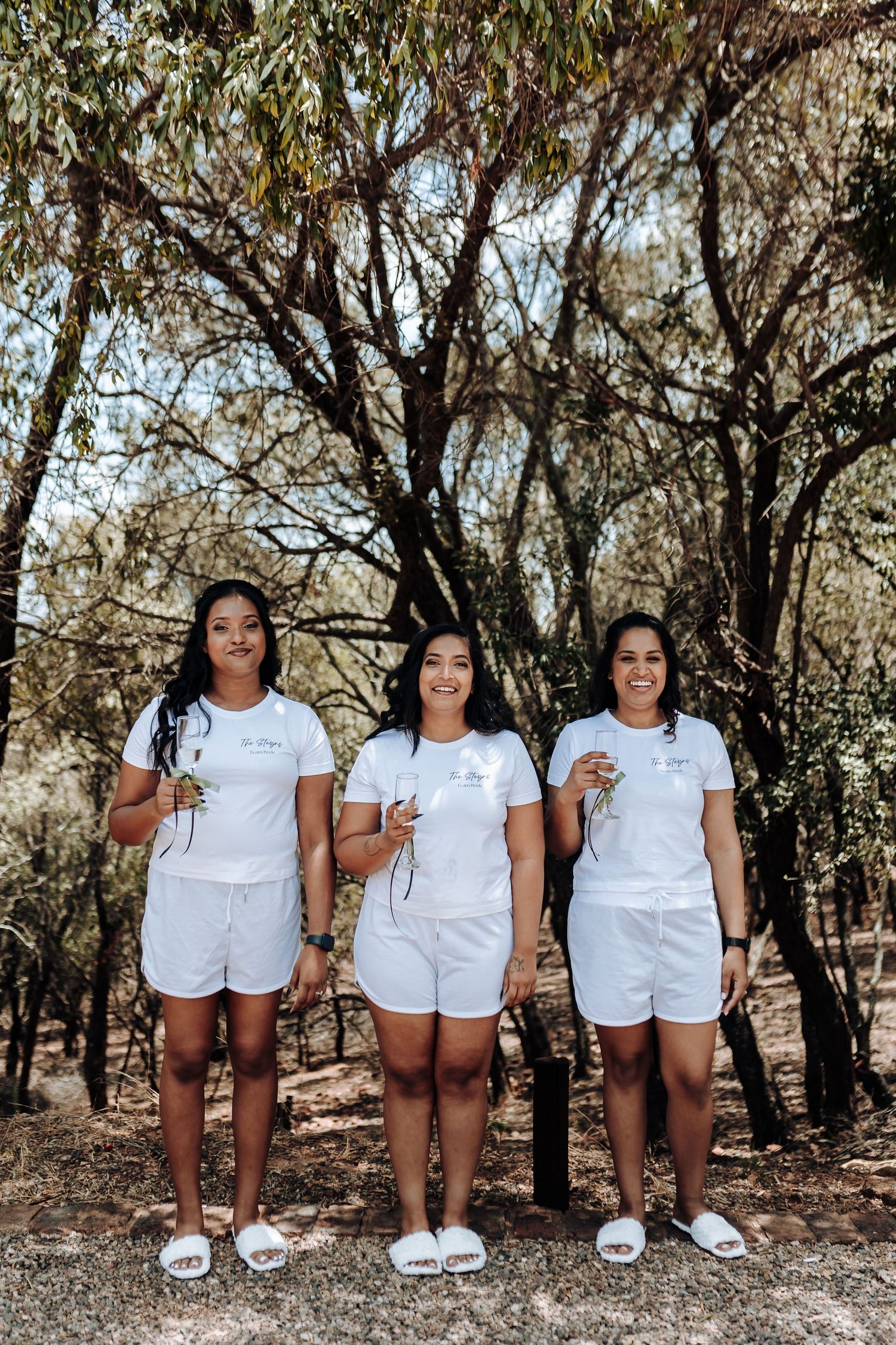 Three people in matching white outfits standing together outdoors among trees.