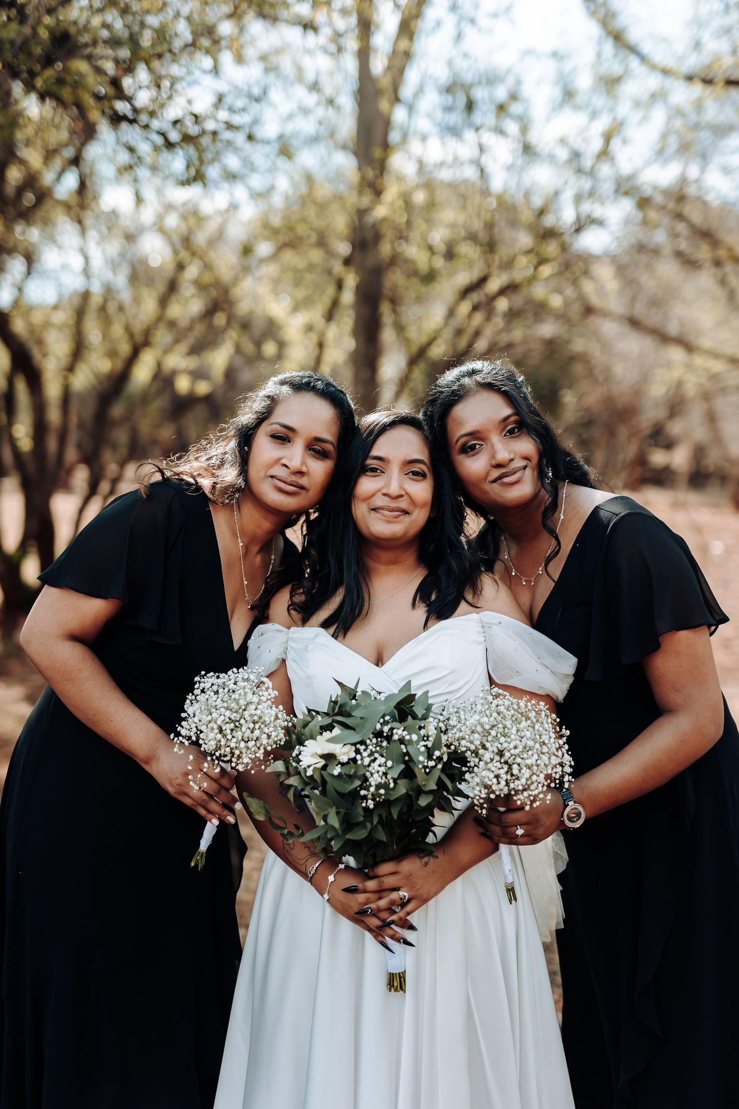Bridesmaids in black dresses embrace bride with white floral bouquets.