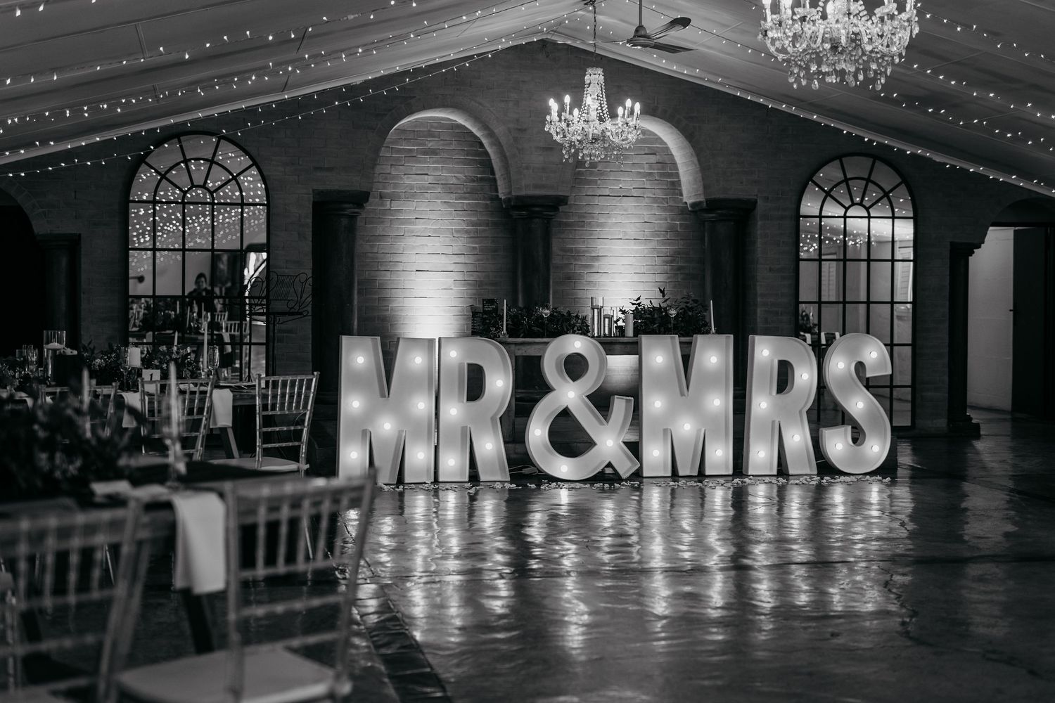 Black and white image of illuminated MR&MRS sign in elegant ballroom with chandeliers.