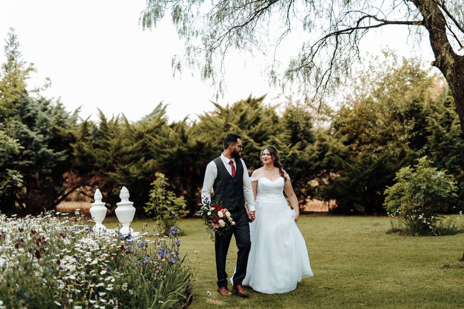 A wedding couple walks together through a garden filled with white flowers and evergreen trees.