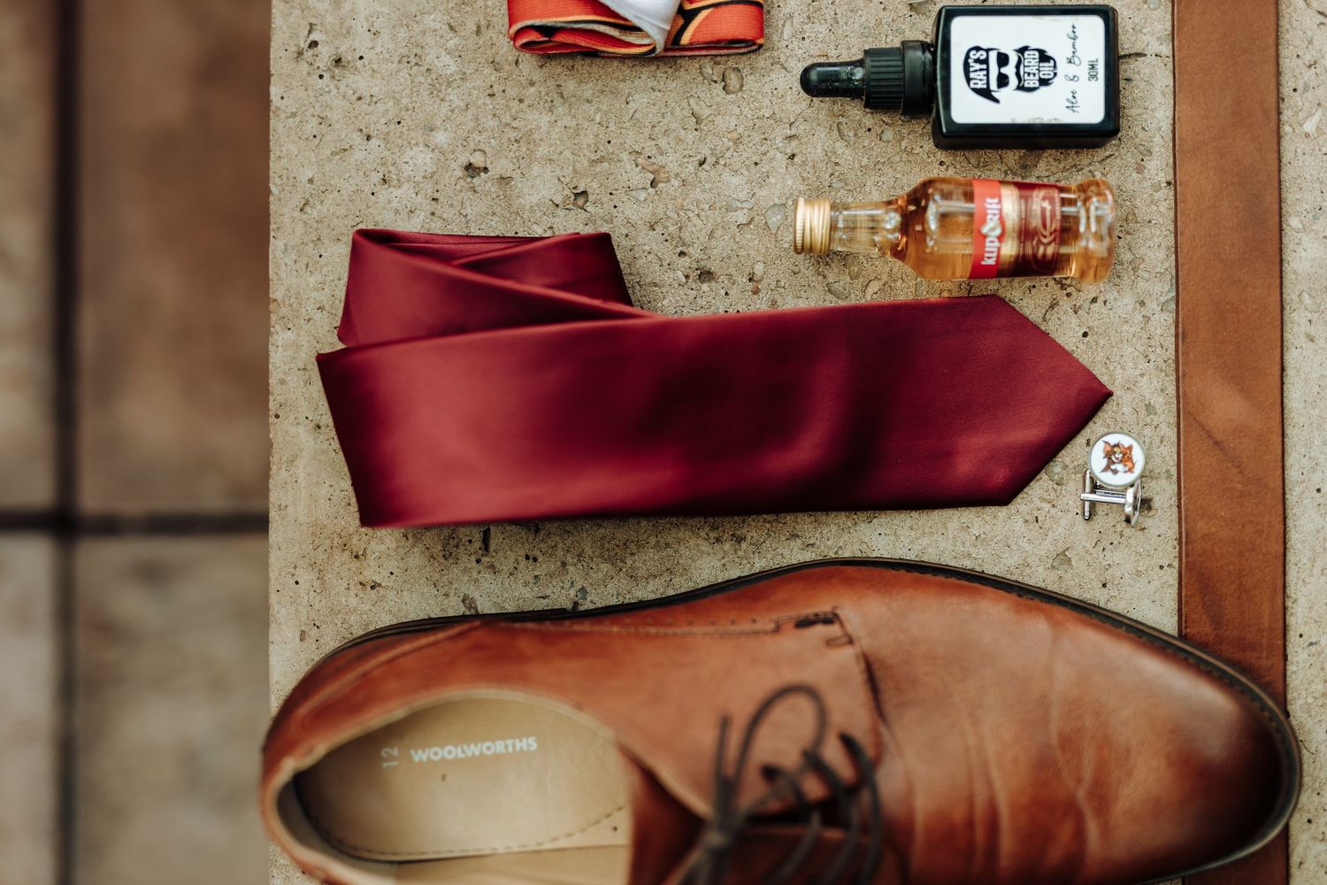 Wedding accessories featuring brown leather shoes and burgundy tie on concrete surface.