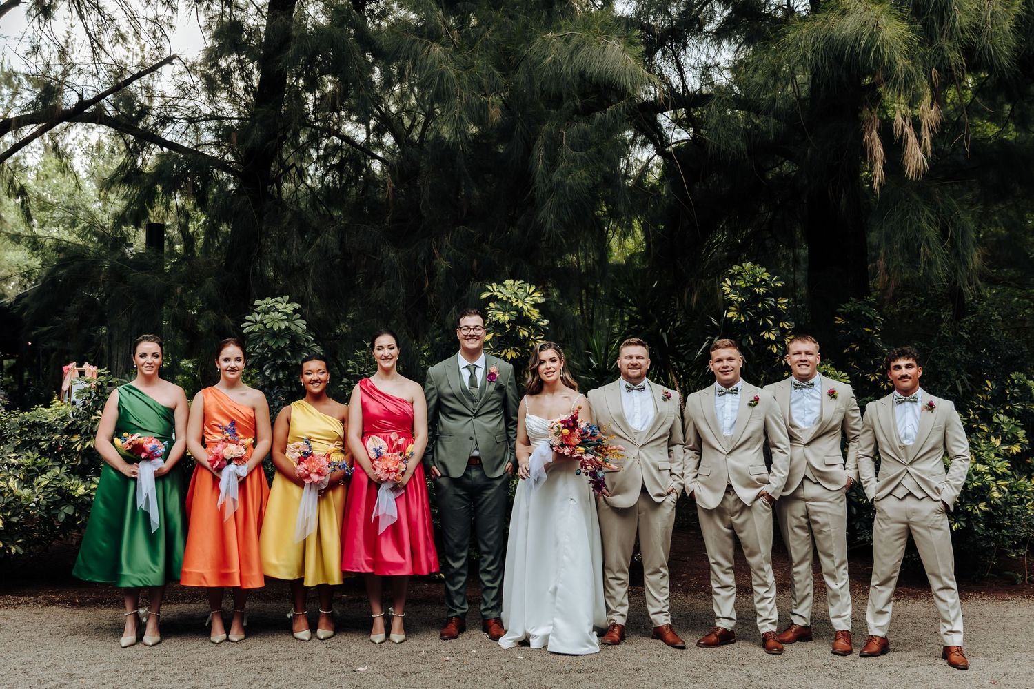 Wedding party lined up outdoors showing bridesmaids in bright rainbow colored dresses and groomsmen in beige suits.