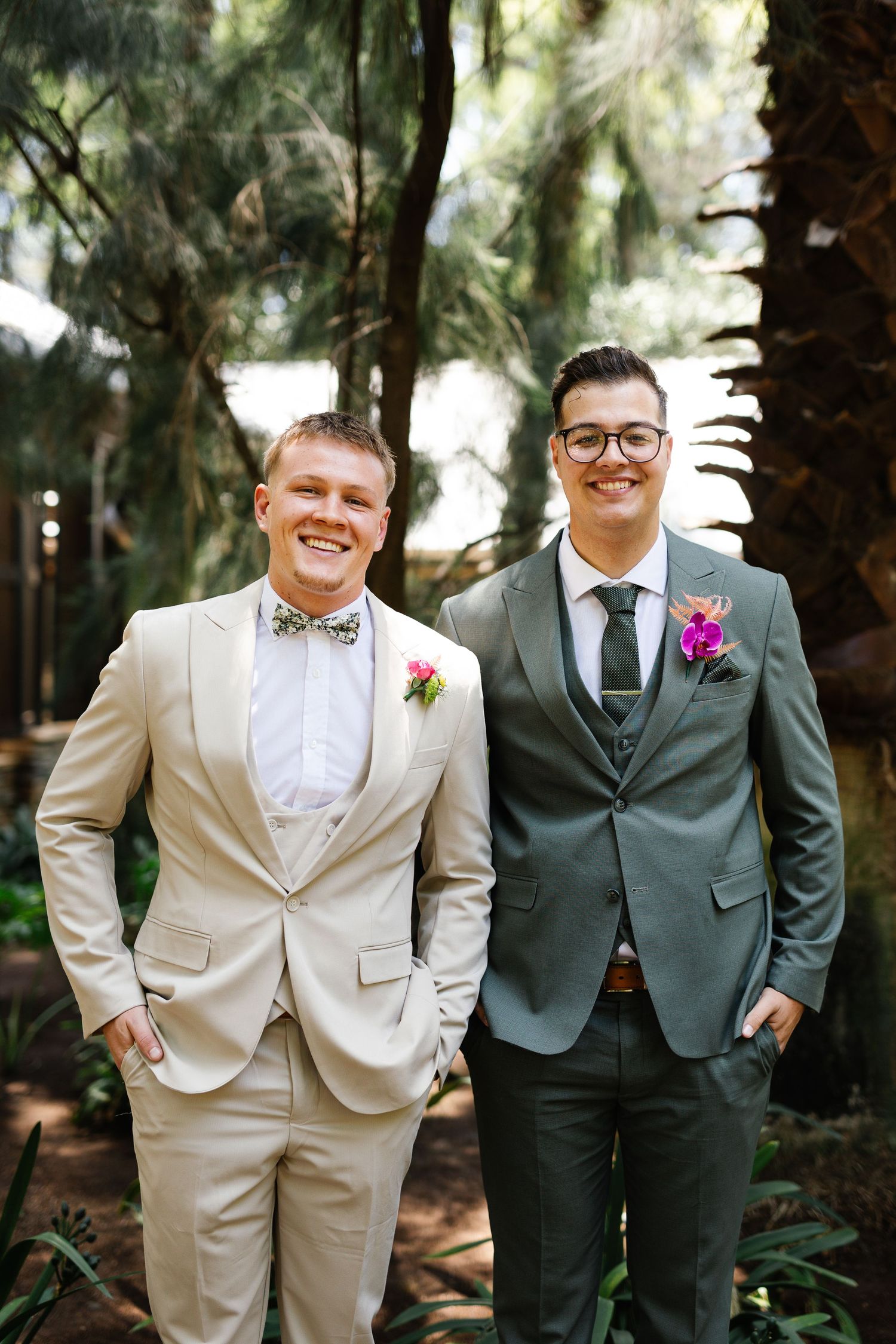 Two people in contrasting cream and sage suits stand together in a forest setting during a wedding.
