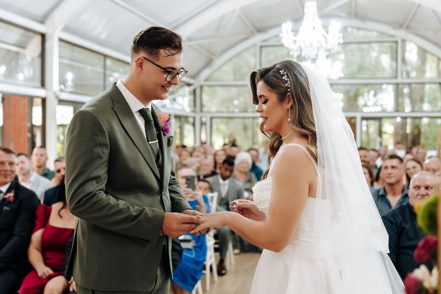 A couple exchanging rings during their wedding ceremony in a sunlit glass-roofed venue.