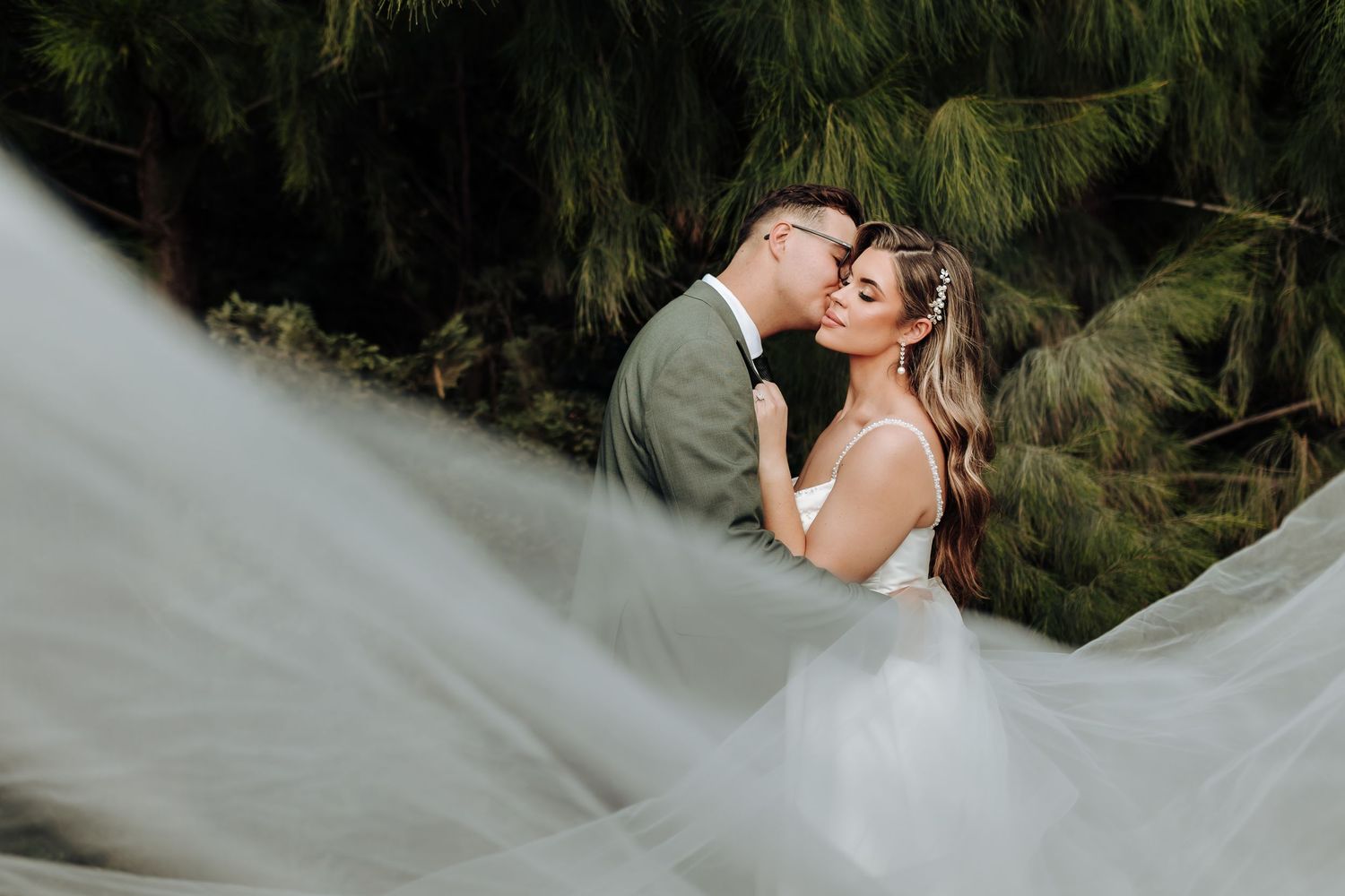 A couple shares a romantic kiss in a forest setting with flowing white wedding dress fabric creating ethereal movement.