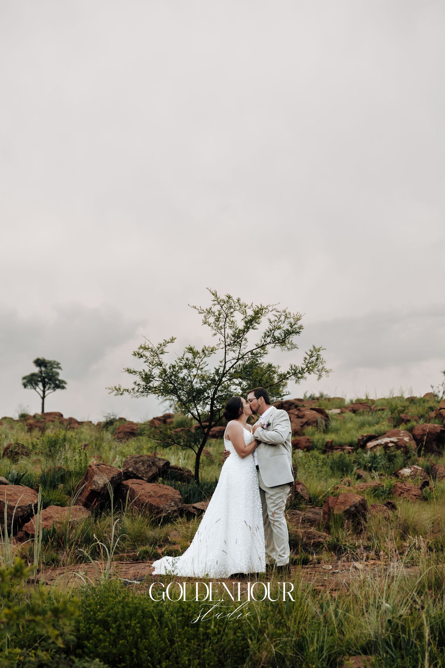 A romantic wedding photo session takes place in a rugged desert landscape with rocky terrain and scattered vegetation.