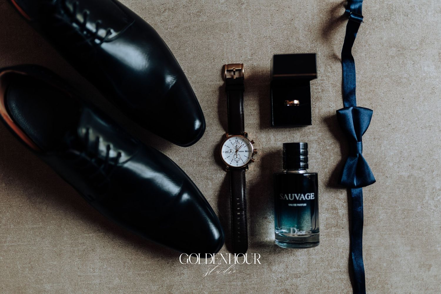 Wedding accessories including black dress shoes, watch, cologne, and blue bow tie arranged on beige surface.