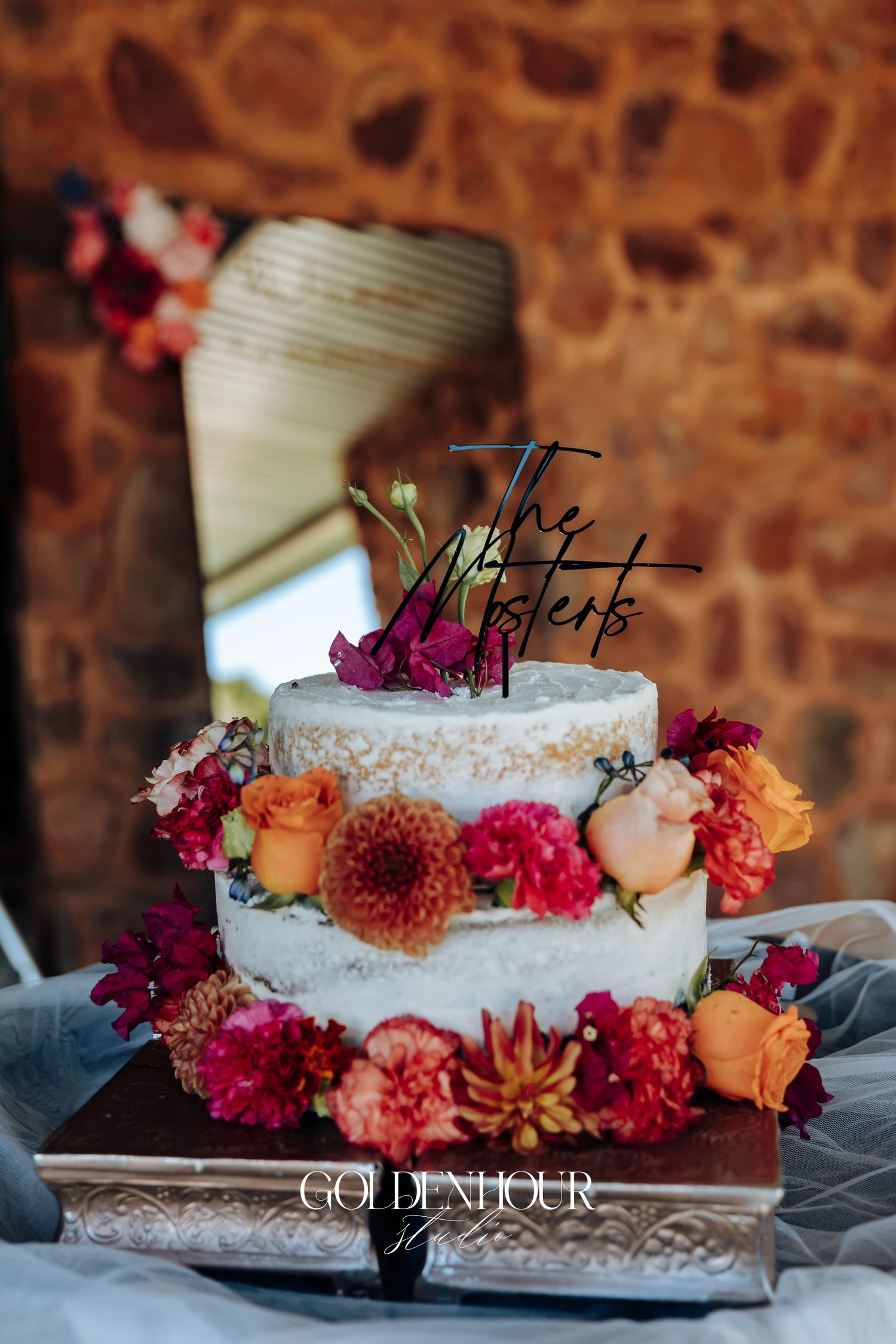 A rustic wedding cake decorated with vibrant orange and pink flowers sits on a lace doily against a stone wall background.