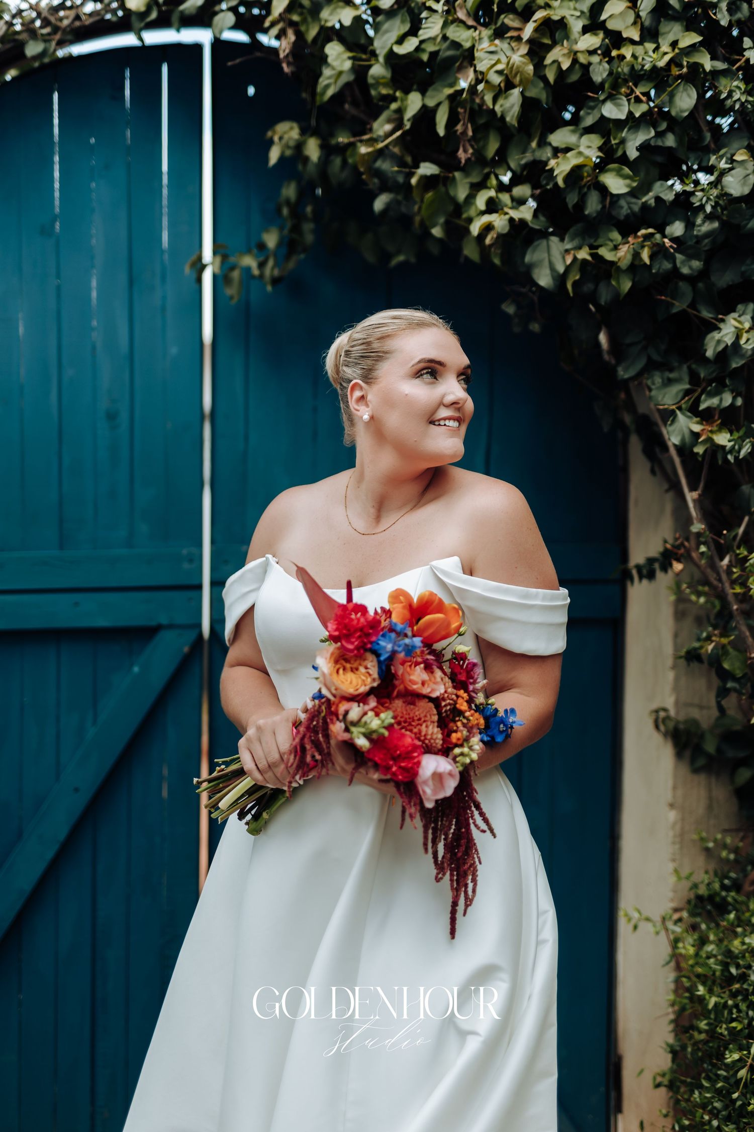 Elegant bride in off-shoulder white gown holds vibrant autumn bouquet with orange and blue flowers against teal door.