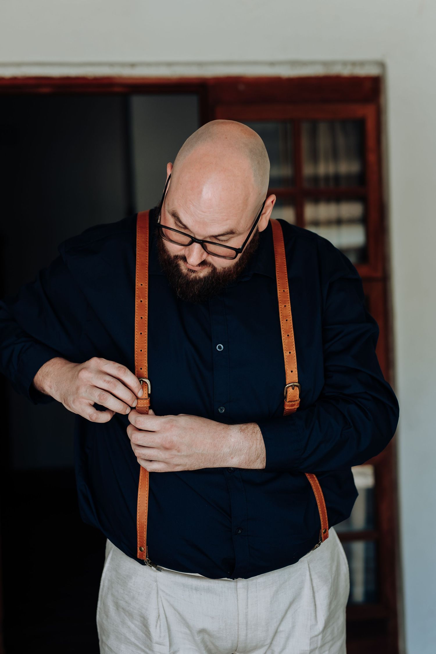 A person in a navy shirt and tan suspenders stands against a dark background with crossed arms and contemplative pose.