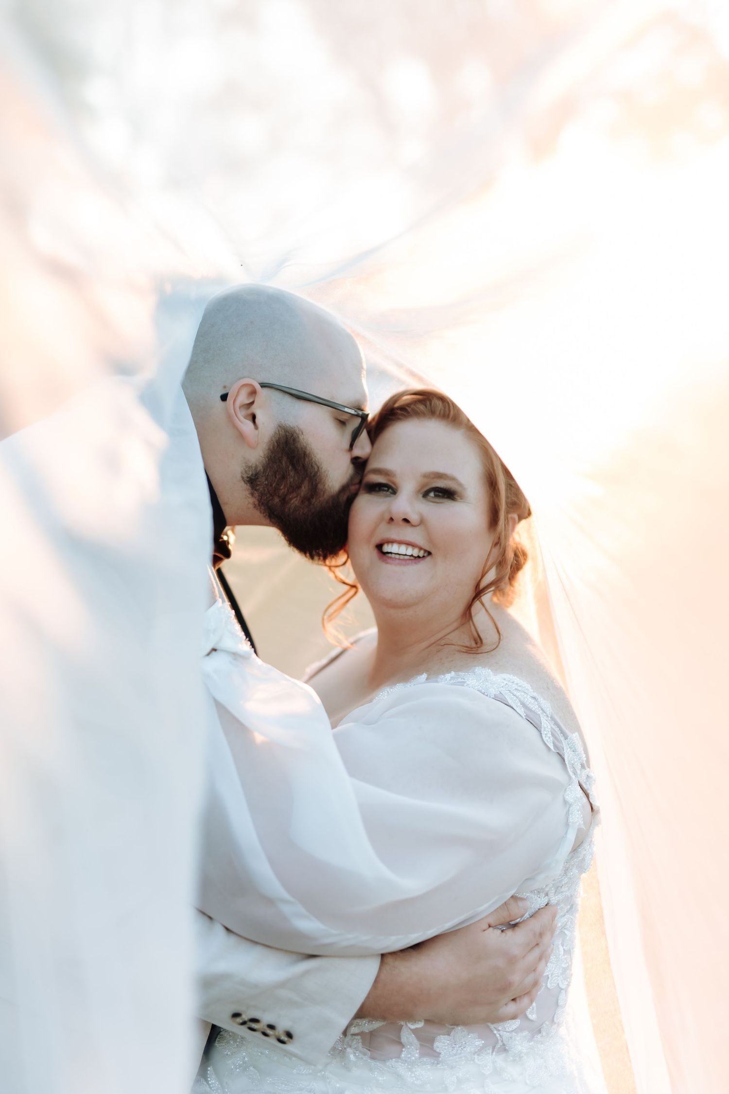 A romantic wedding portrait of a couple embracing in soft sunlight, with the bride wearing a white dress with flowy sleeves.
