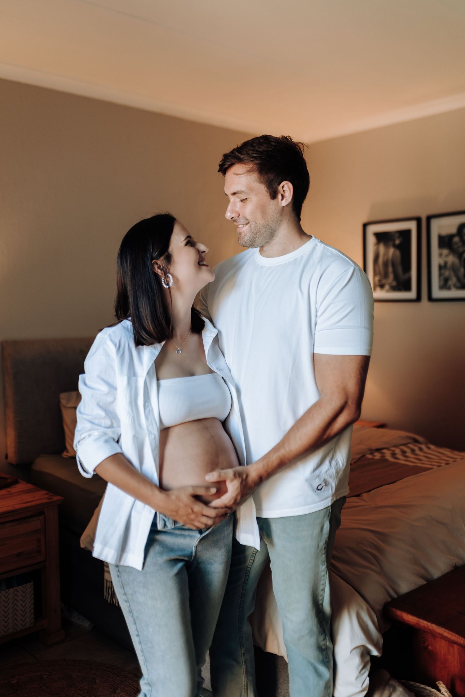A couple wearing white shirts and jeans shares an intimate moment together in a cozy bedroom setting.