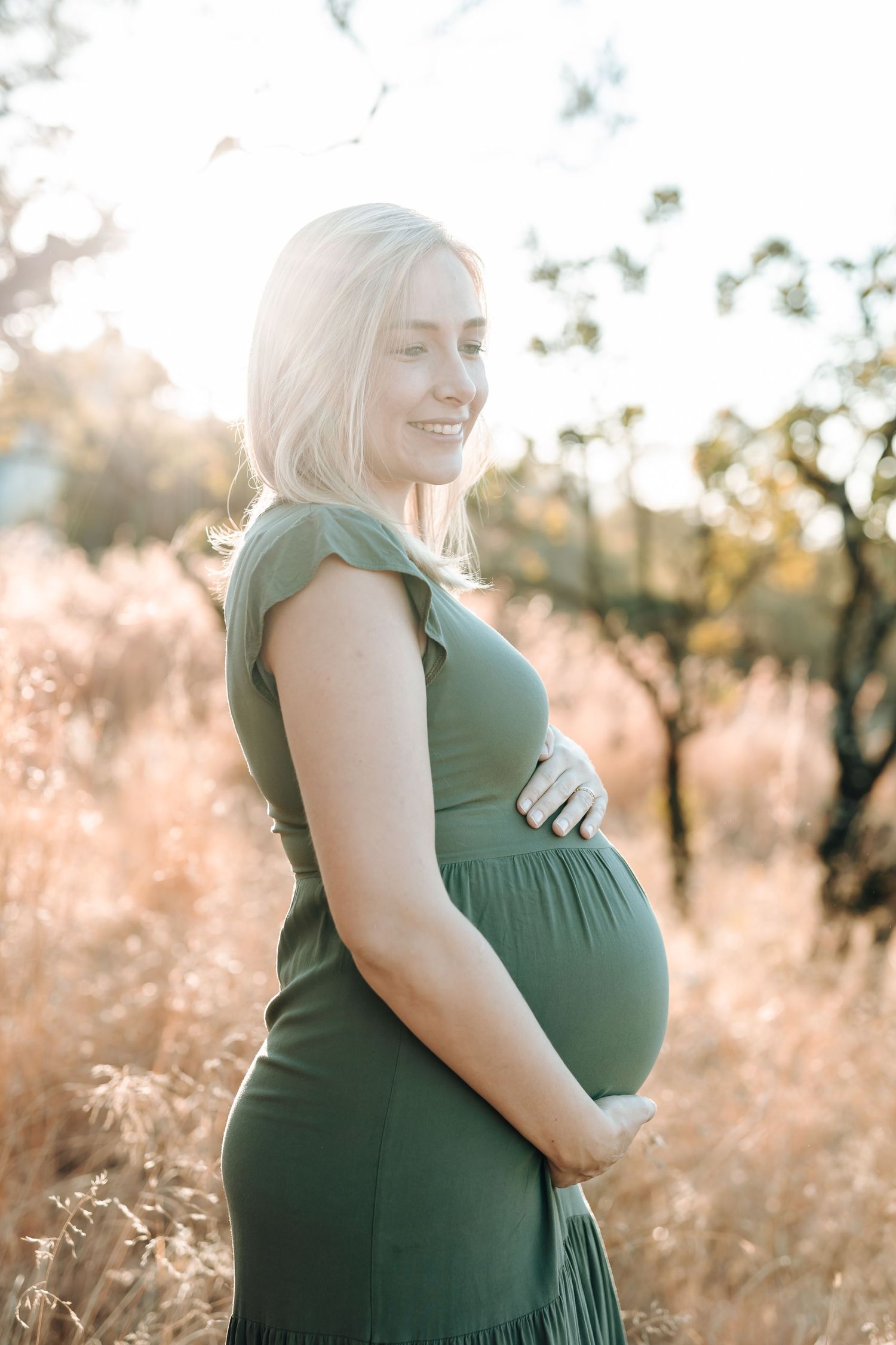 Person in green maternity dress poses in golden sunlit field with tall grass during sunset.