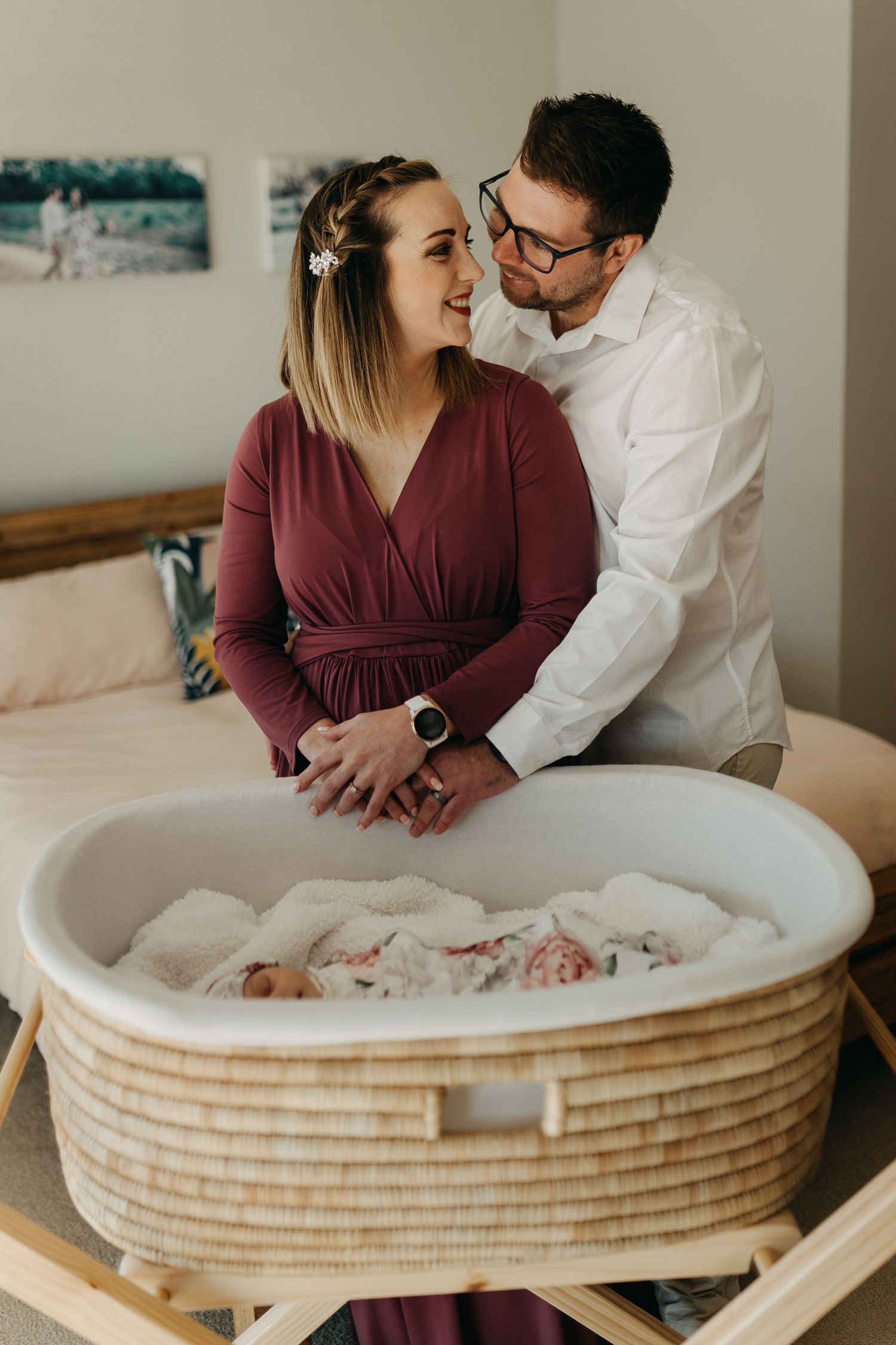 Couple in burgundy and white looking lovingly at newborn in woven bassinet.
