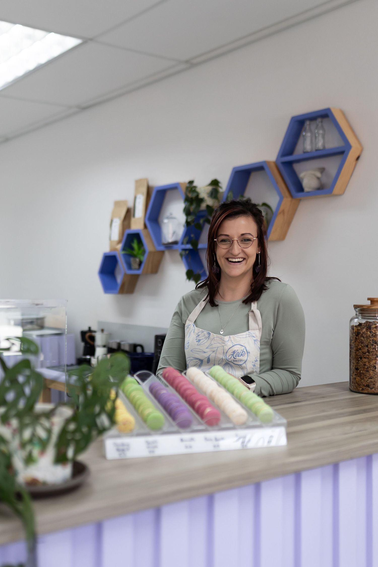 Person smiling and sitting at white desk with hexagonal shelves and plants in office space.