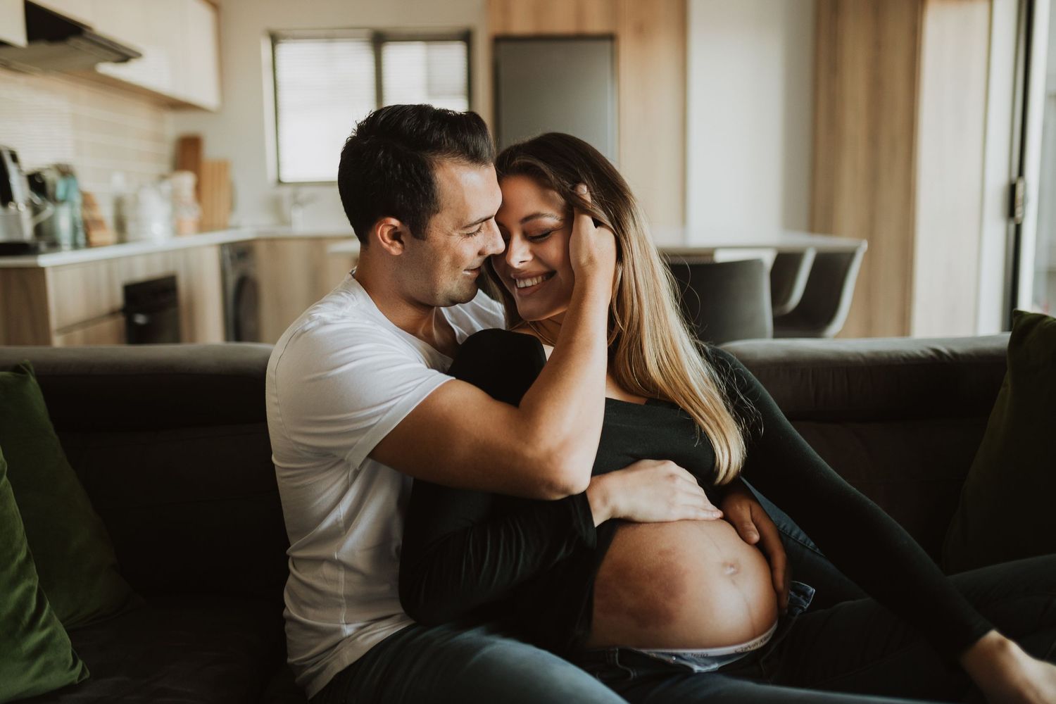 A couple embraces intimately on their couch during a maternity photo session in their living room.