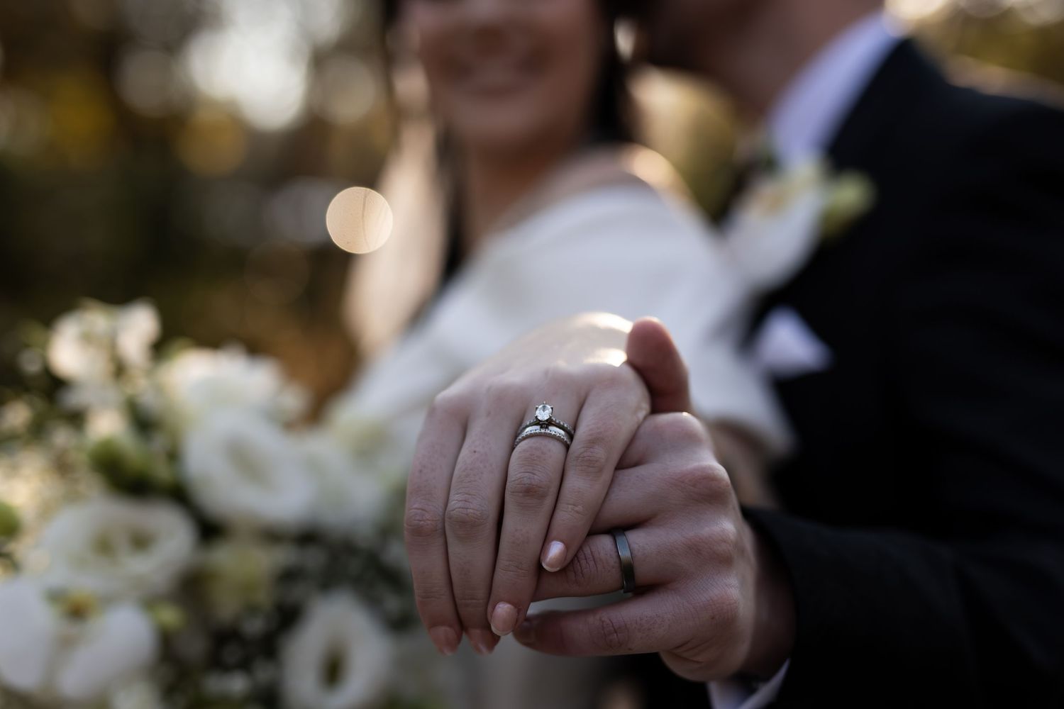 Close-up photo showing clasped hands with a diamond engagement ring glinting in soft focus light outdoors.
