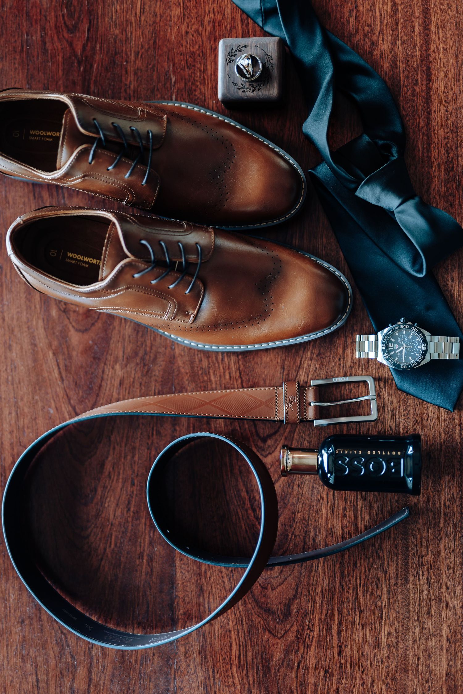 Brown leather dress shoes arranged with a teal tie, leather belt, watch and cologne bottle on dark wooden surface.