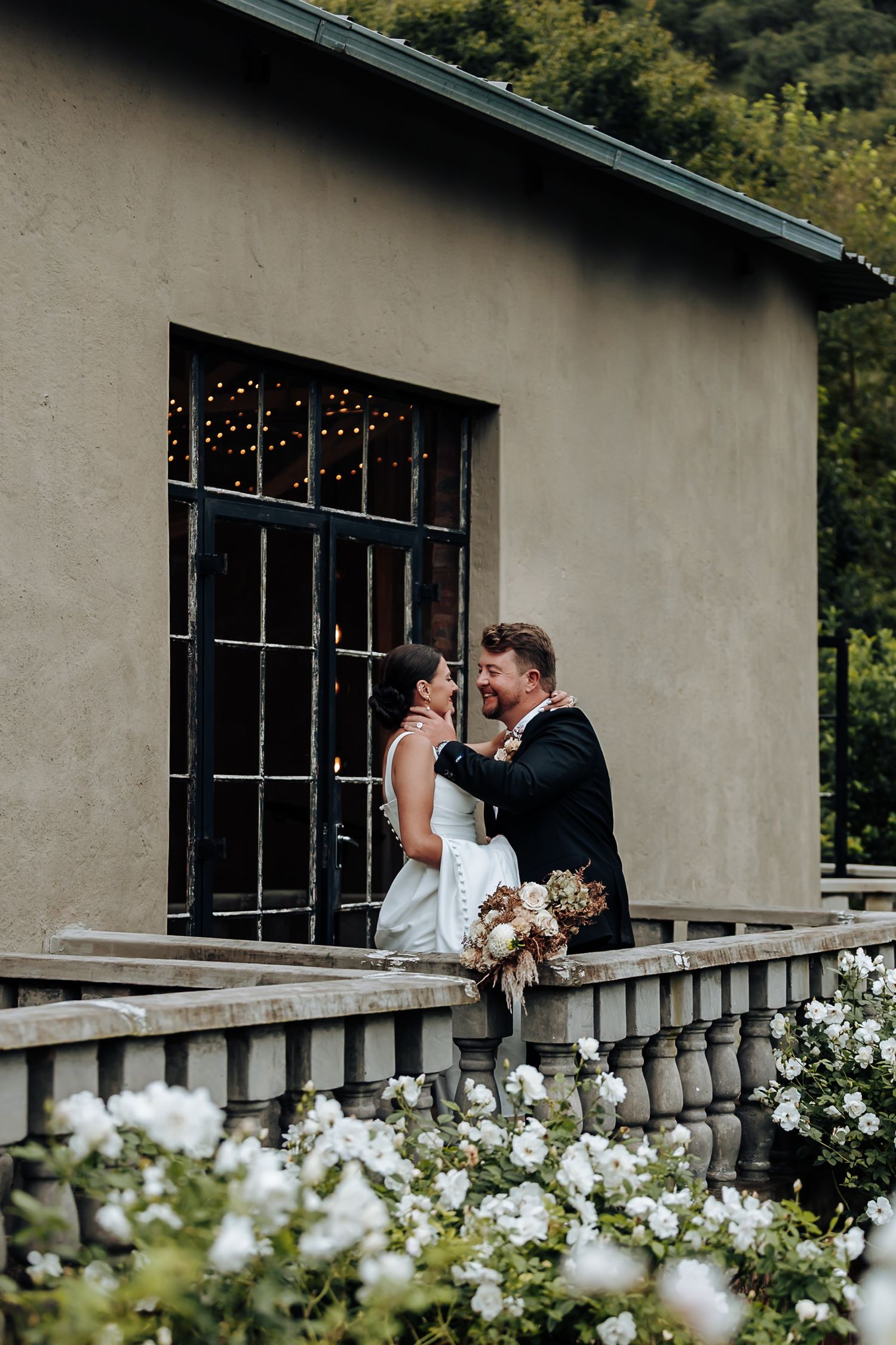 A romantic moment shared between a couple on a balcony surrounded by white roses and greenery at a wedding venue.