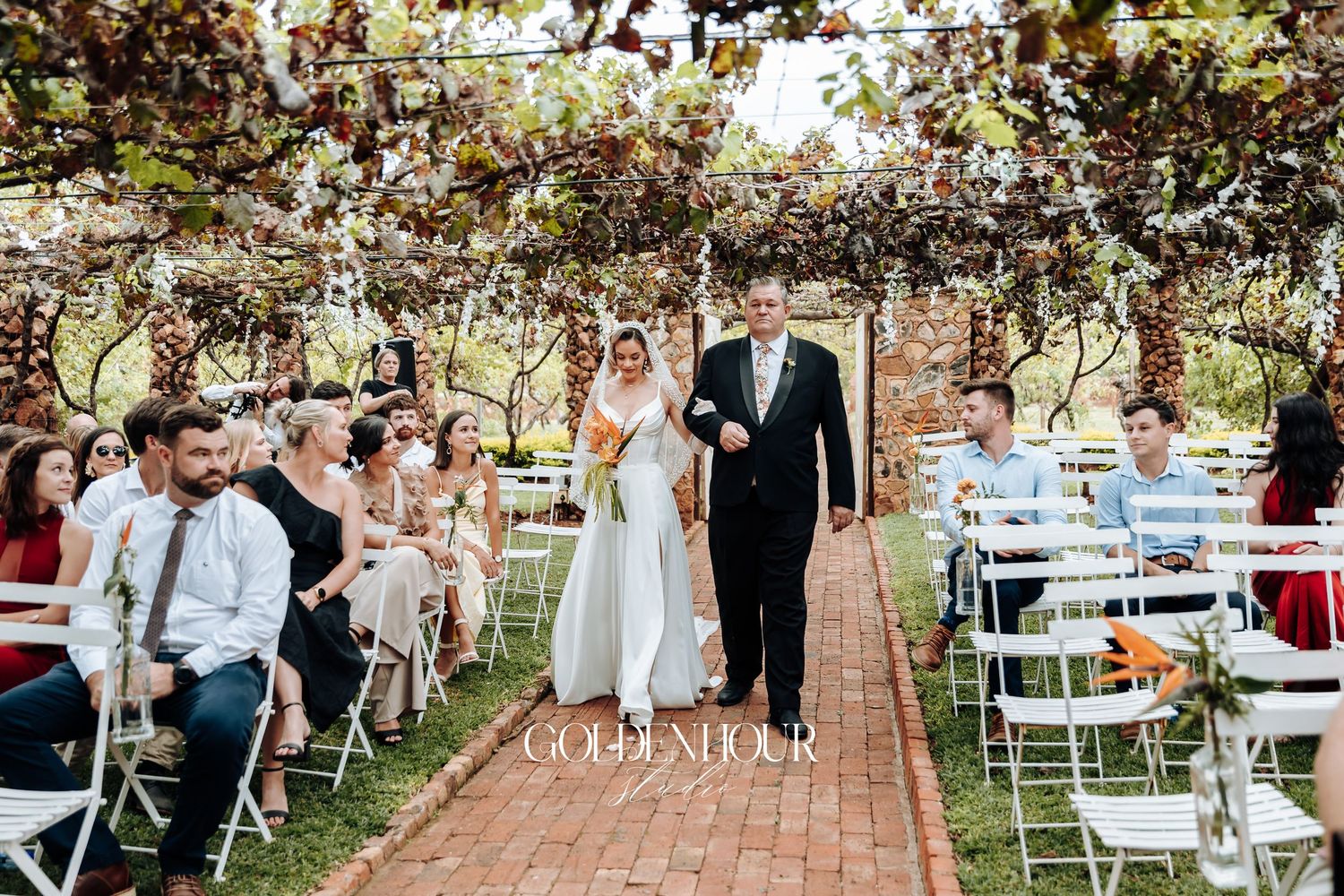 Outdoor wedding reception setup with white chairs and tables under trees.