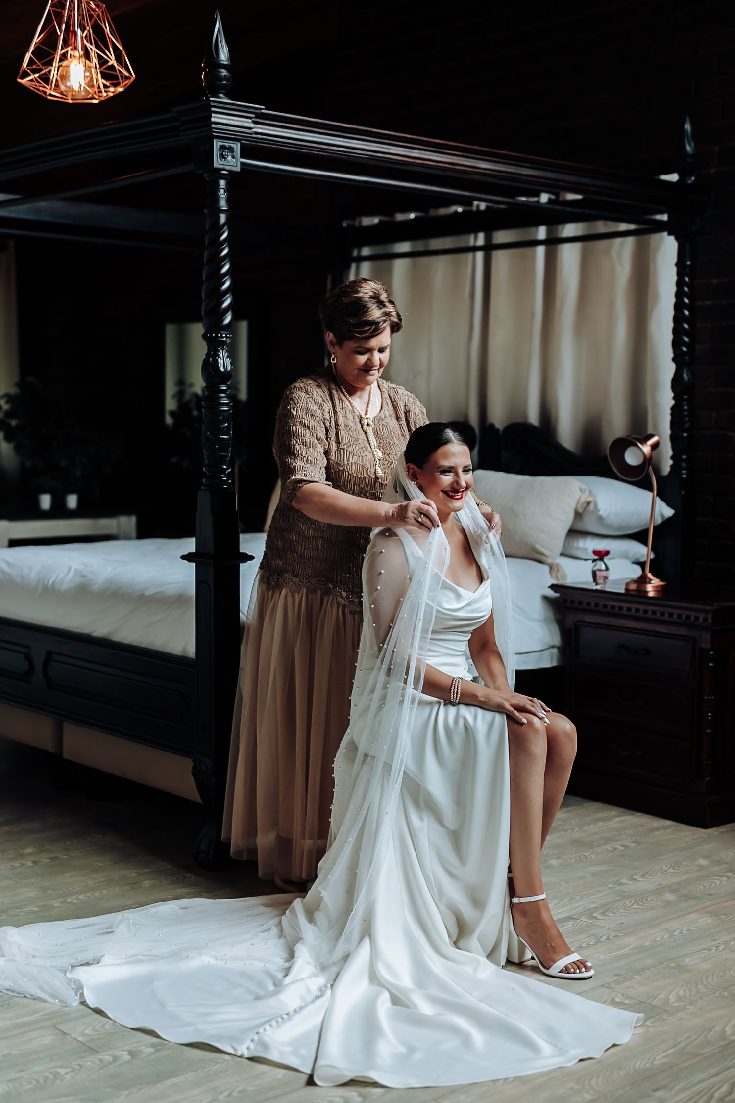 Someone helps adjust a flowing white wedding dress on a person seated in an elegant room with dark columns.