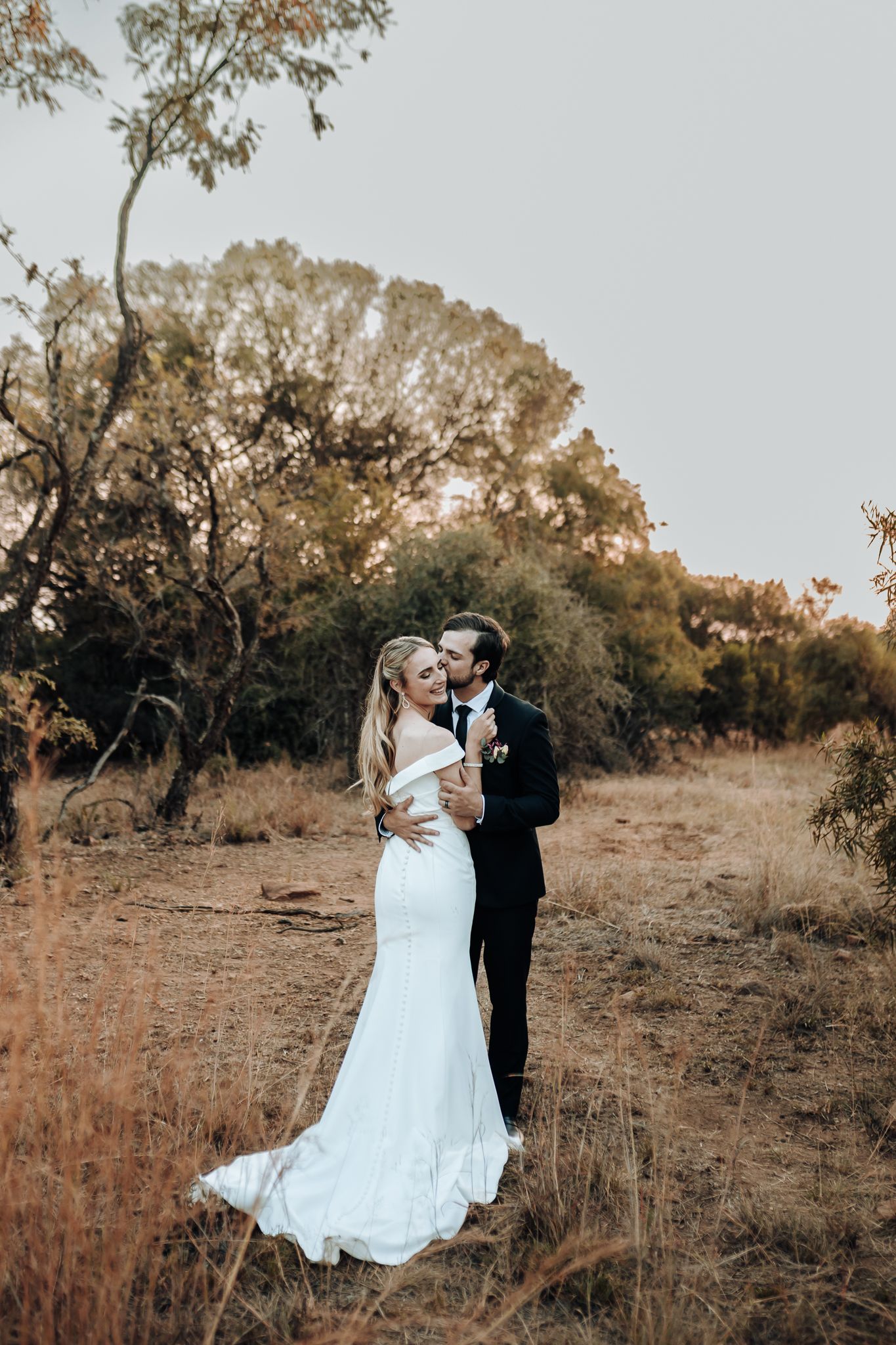 A couple embraces on a rural path surrounded by trees at sunset.