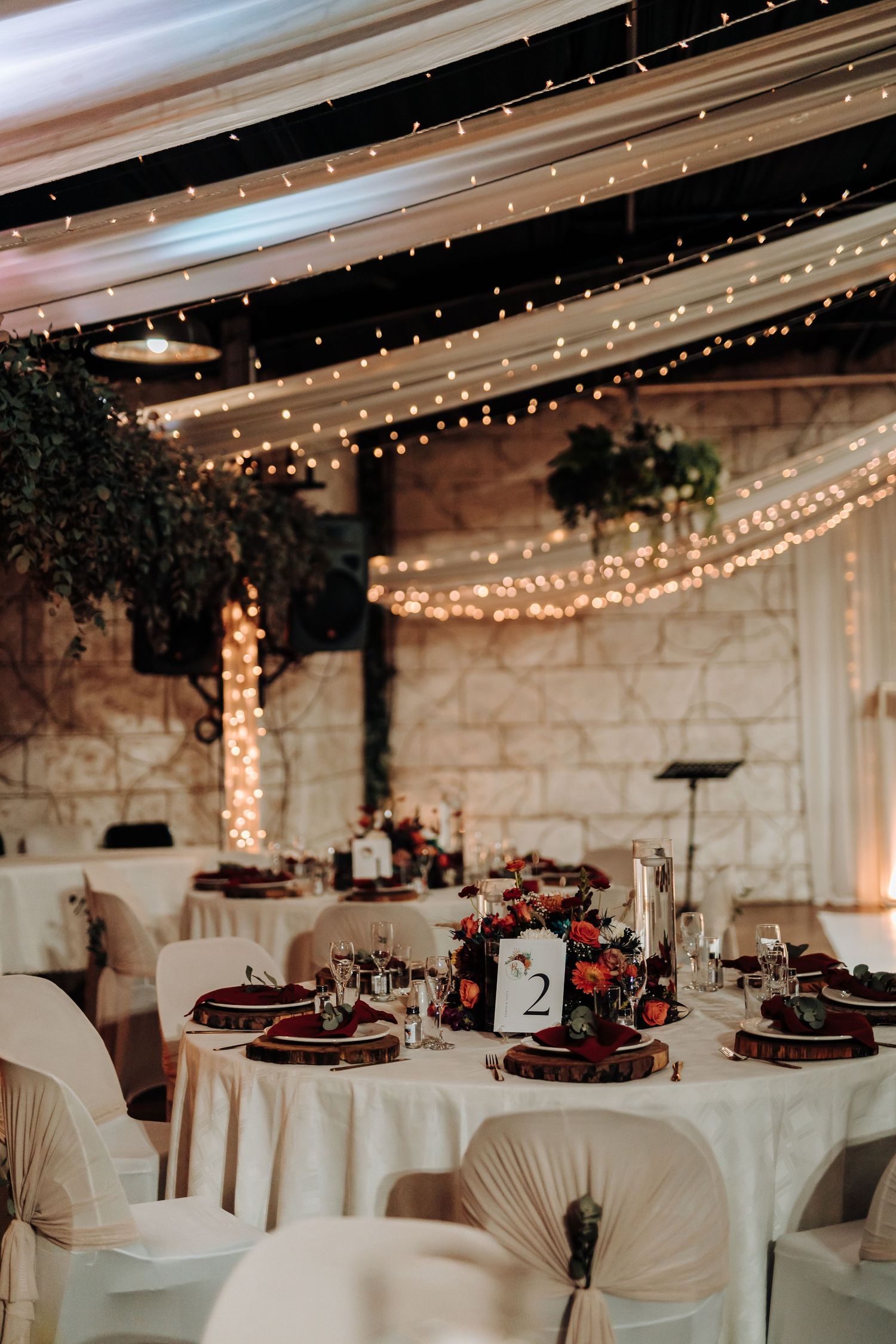 Elegant wedding reception tables with white linens, burgundy accents, and twinkling string lights overhead.