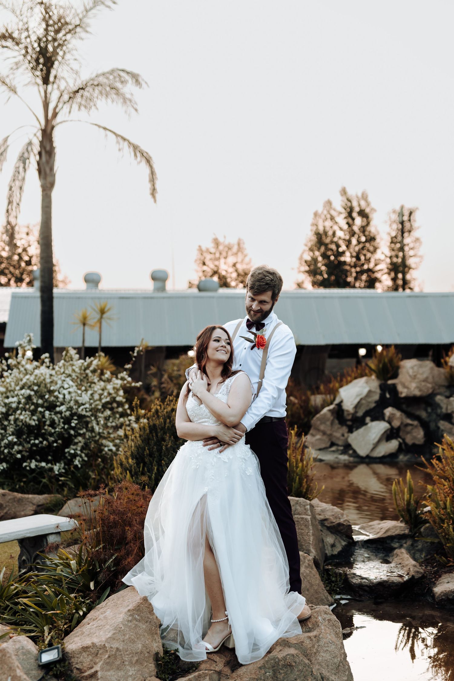 Couple in wedding attire embrace while standing on decorative rocks near palm trees during sunset.