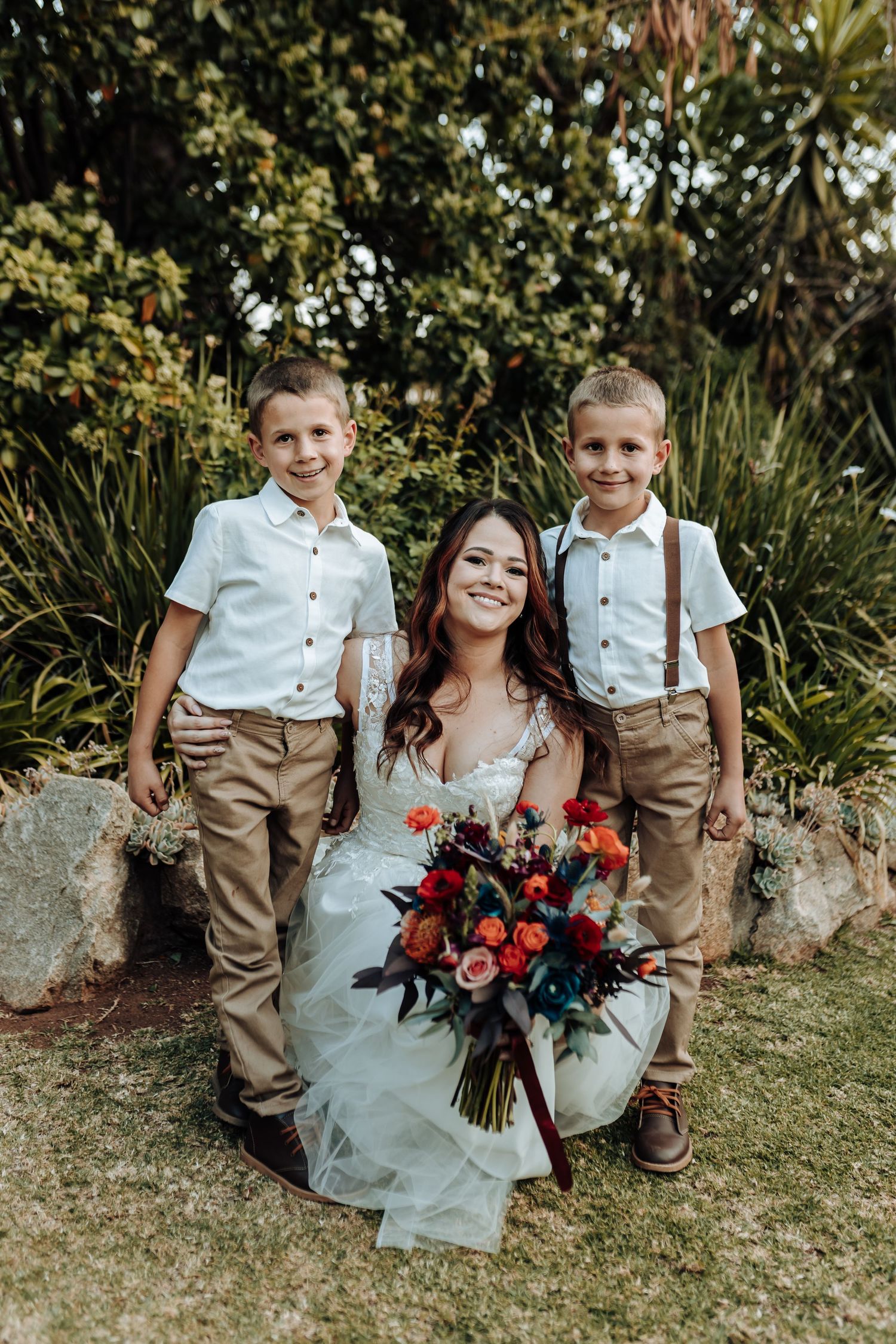 Wedding portrait of bride with young ring bearers in white shirts and suspenders outdoors.