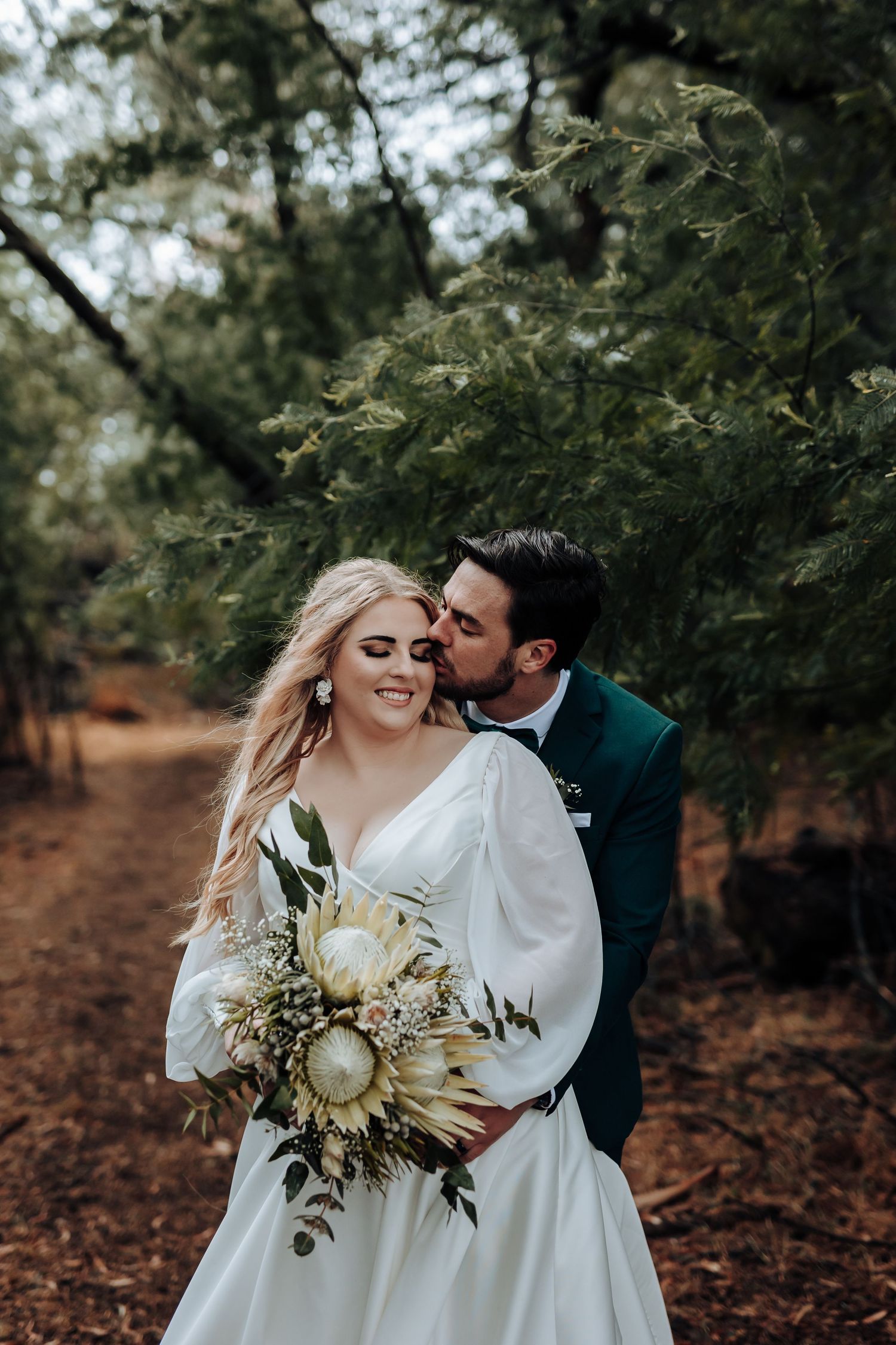 A romantic wedding moment captured in a wooded setting with a stunning dried palm frond bouquet.