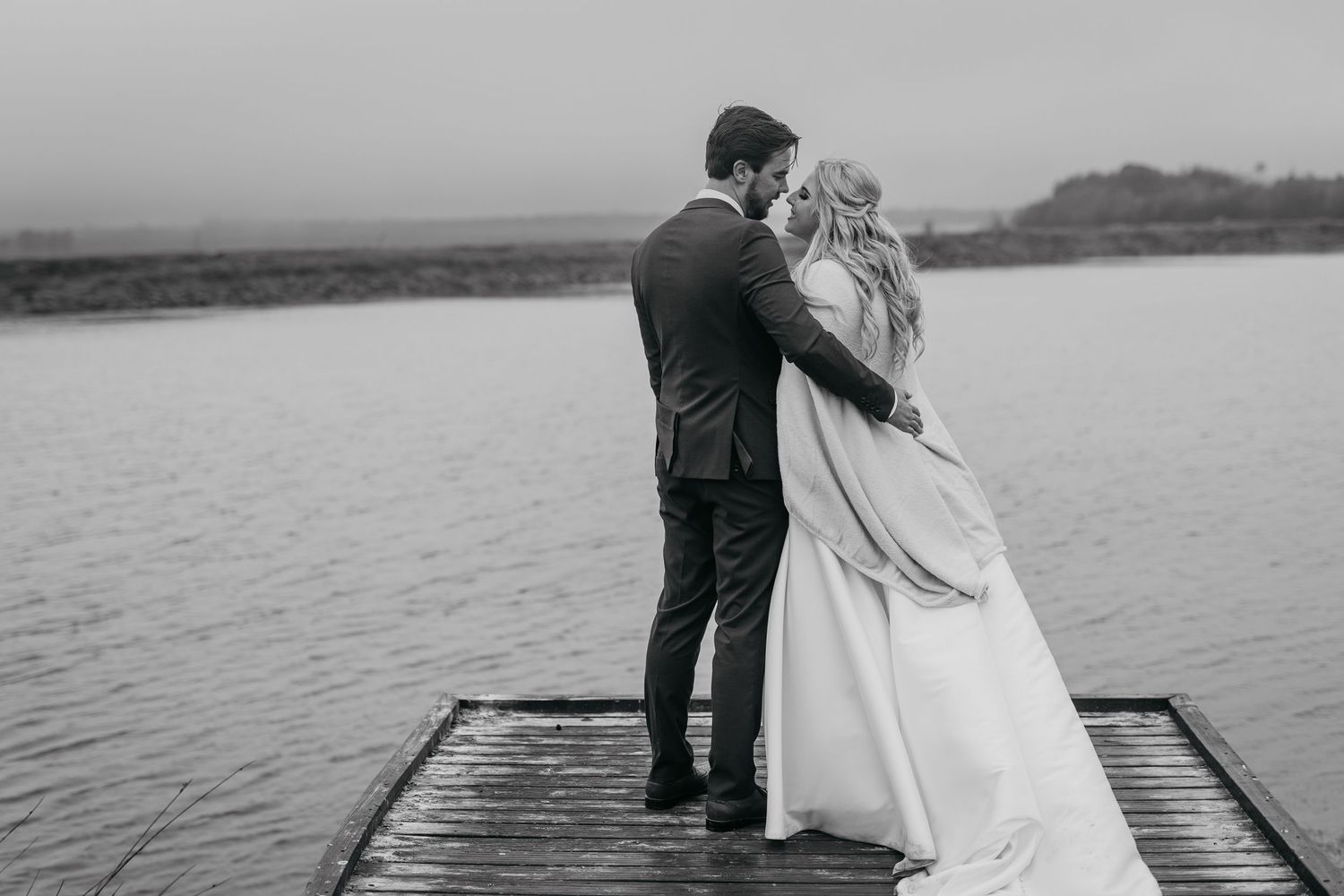 Black and white photo of couple embracing on wooden dock by the water.