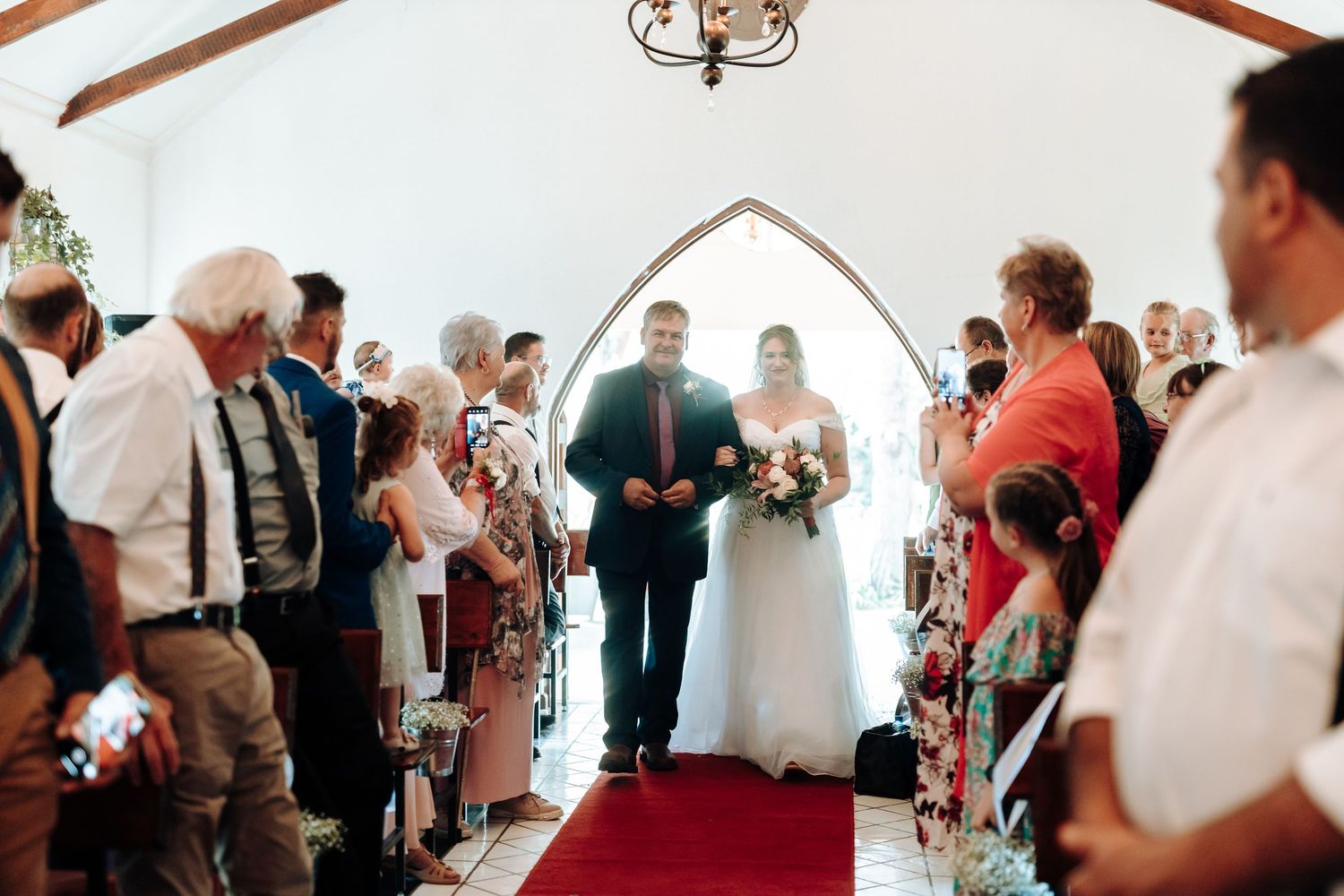 Wedding ceremony procession down red carpet aisle in white chapel with exposed beams.