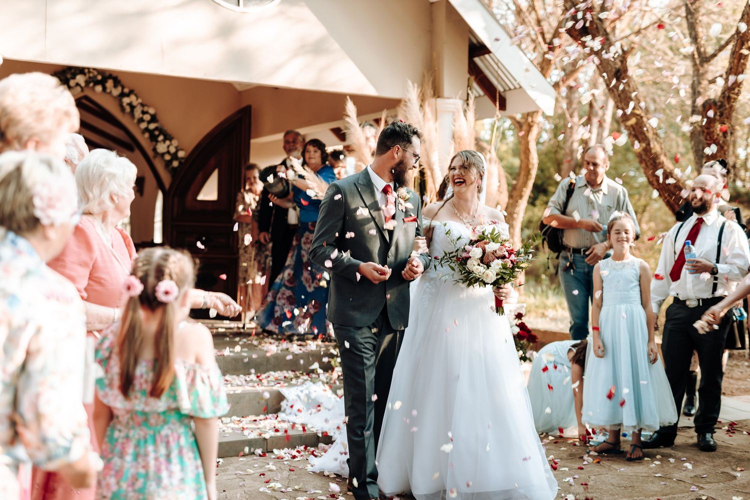 Wedding guests toss confetti as couple exits venue under sunny skies.