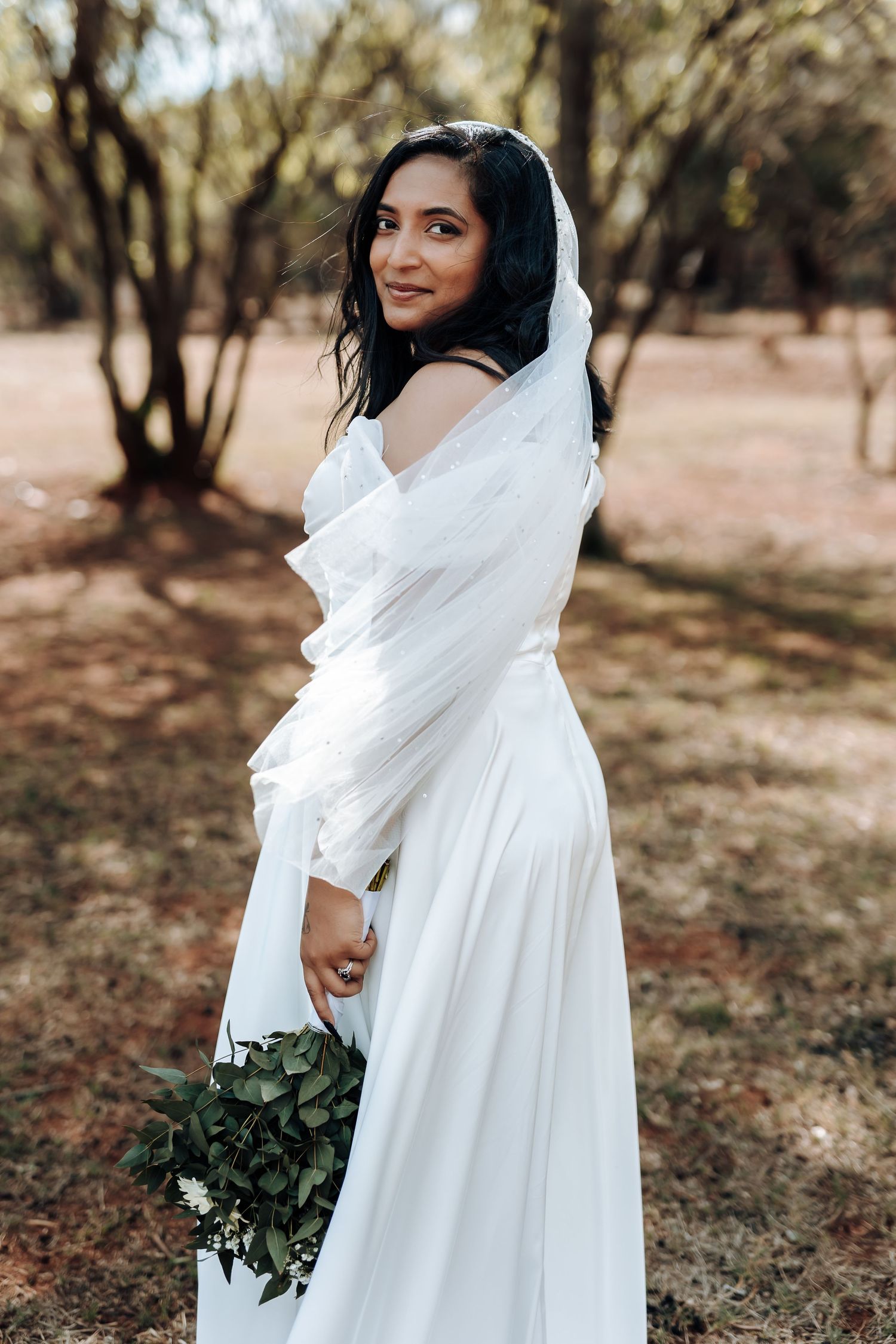 Bride in flowing white dress poses with greenery bouquet in outdoor setting.
