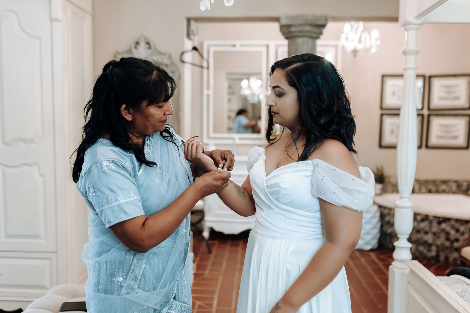 Two people in a bridal preparation room, one helping adjust the other's dress.