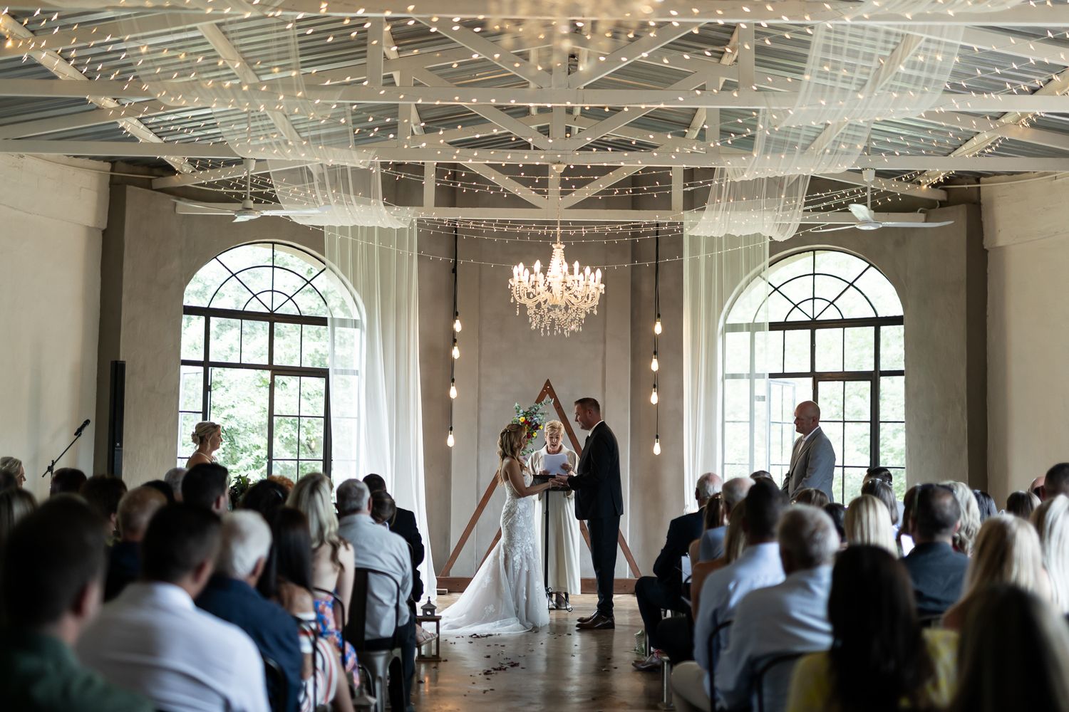 An elegant wedding ceremony takes place in a bright venue with chandeliers and arched windows.