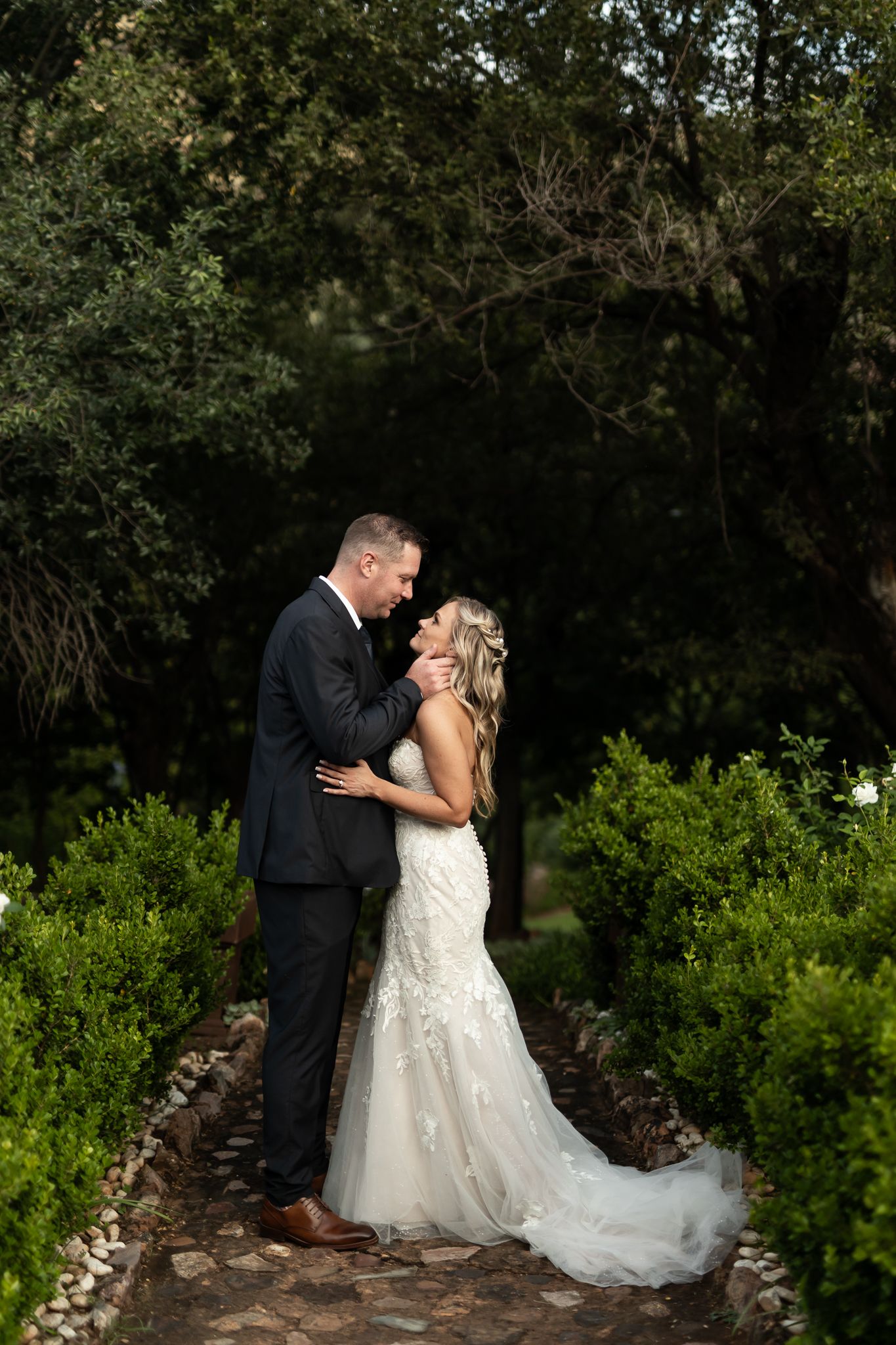 A romantic wedding couple embraces on a garden path surrounded by lush greenery and trees.