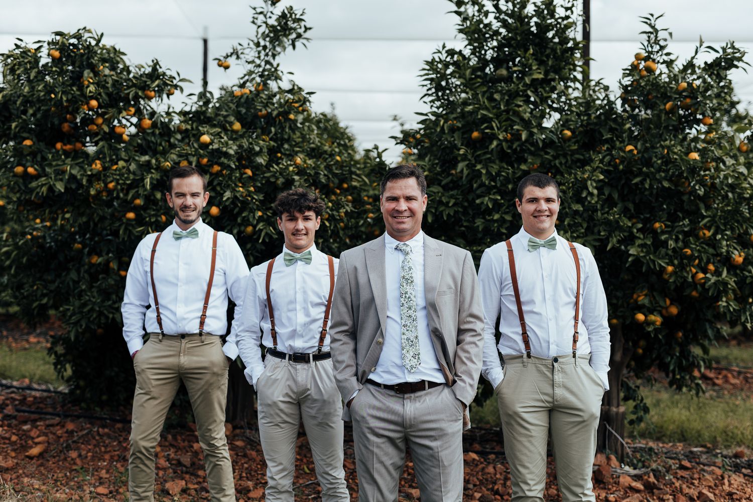 Wedding party in light colored suits with suspenders standing in orange grove.