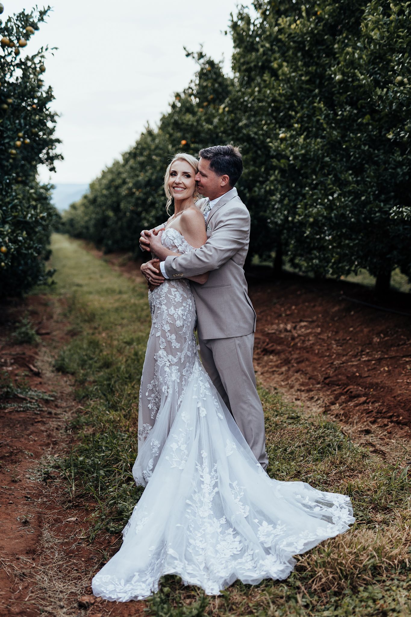 Romantic embrace between newlyweds in orchard with mountains in background.